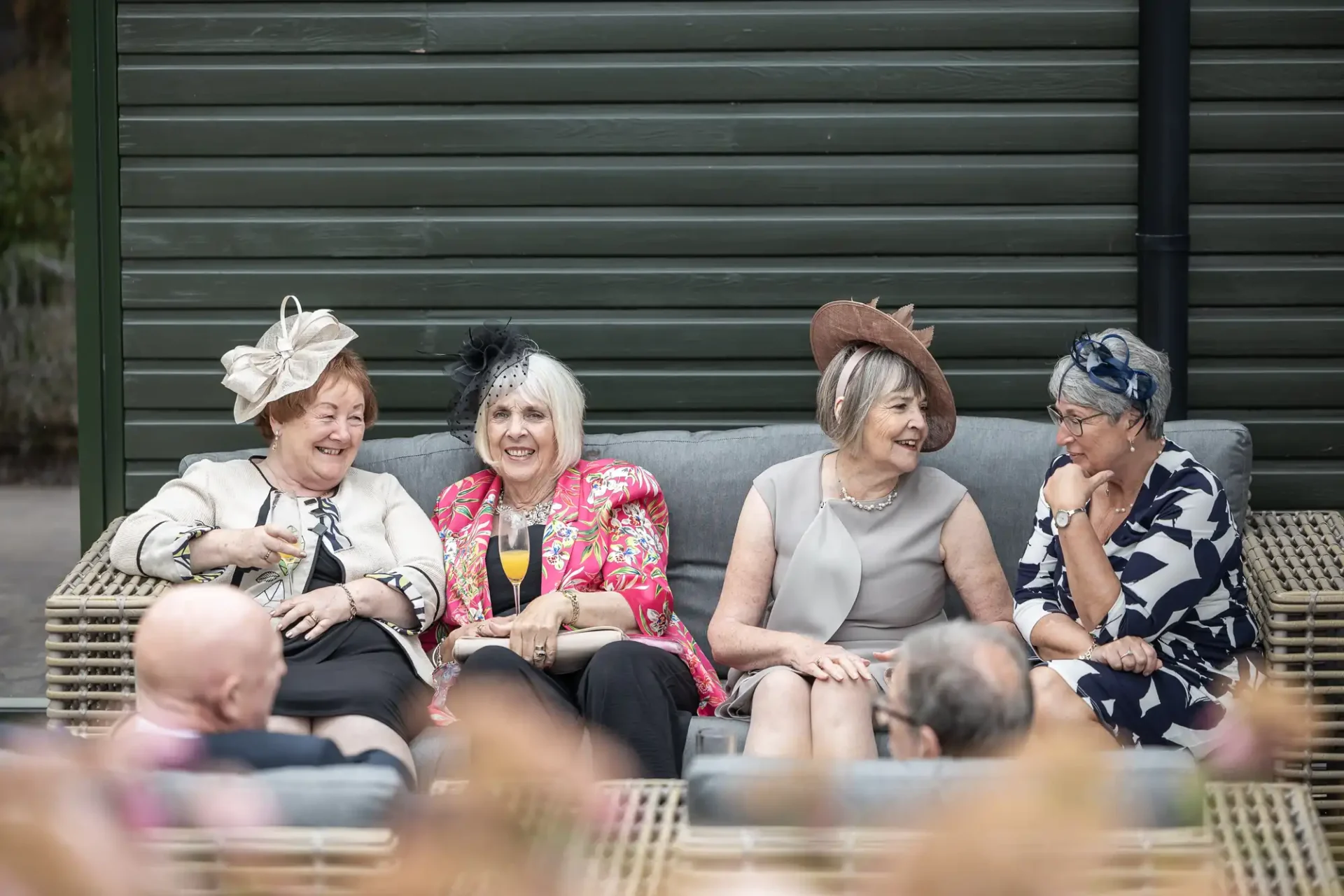 Four women in formal attire and decorative hats sit on an outdoor sofa, chatting and smiling.