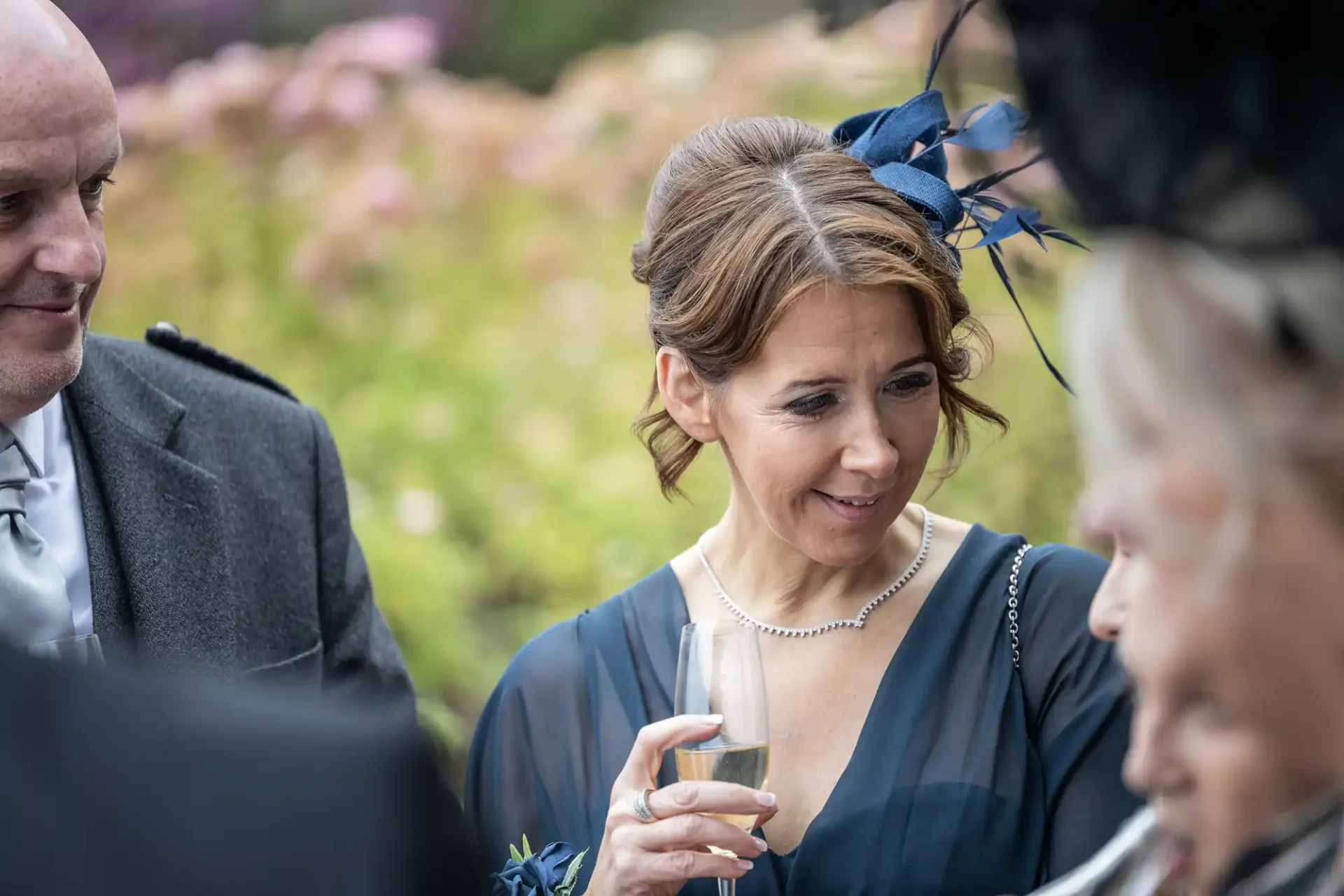 A woman in a blue dress holds a champagne glass and smiles, engaging with others at an outdoor event. She wears a blue fascinator and a pearl necklace. Other people are partially visible.