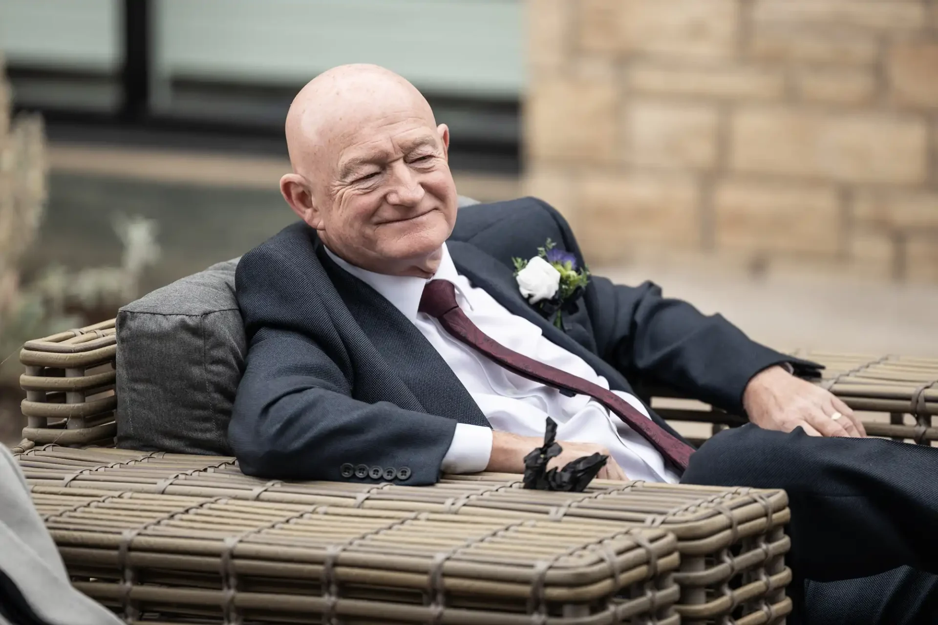 An older man in a suit and tie, with a boutonniere, sits on a wicker chair, smiling slightly.