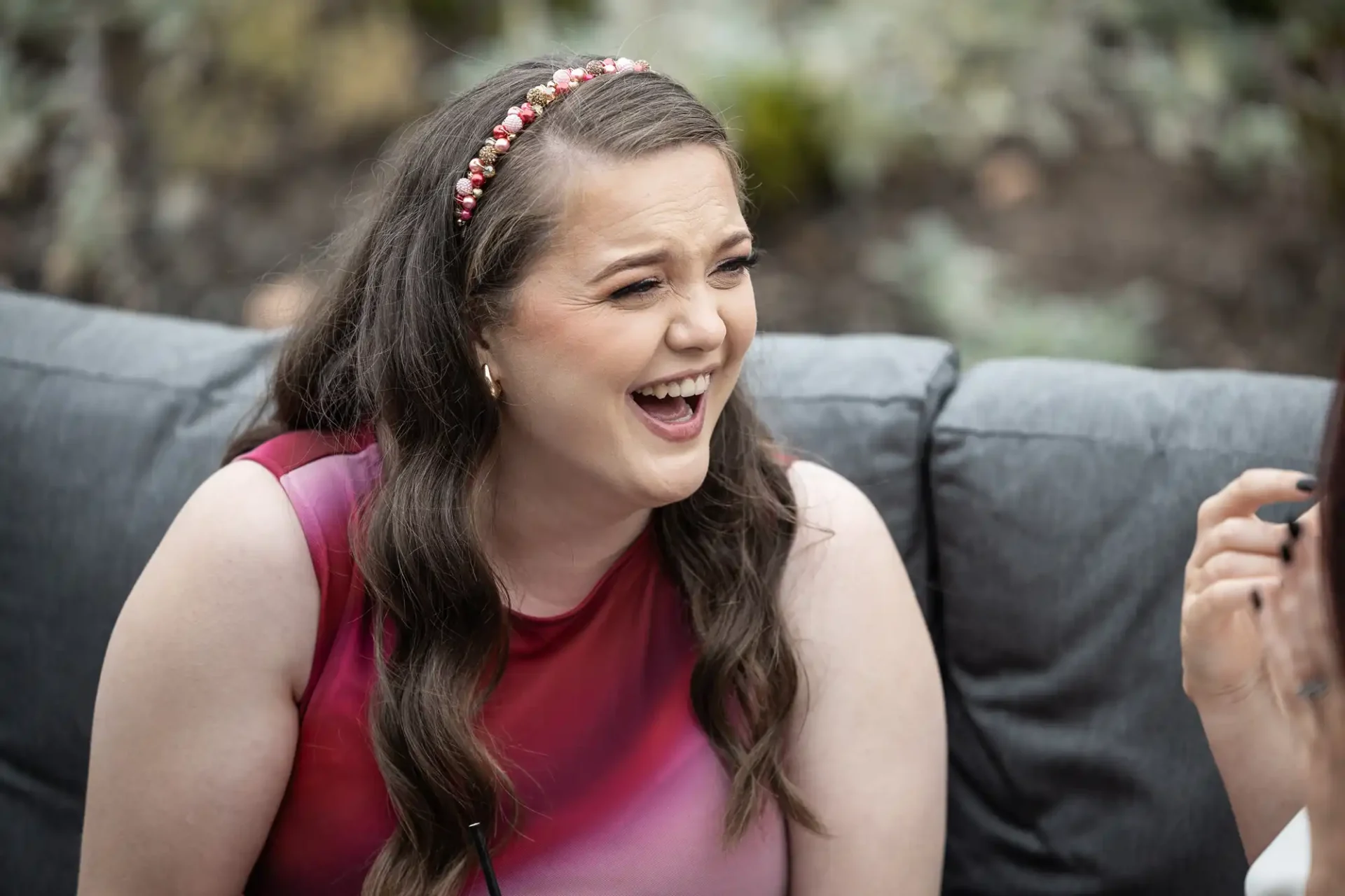 Person with long hair and a headband, wearing a sleeveless top, laughing while sitting on a gray couch outdoors.