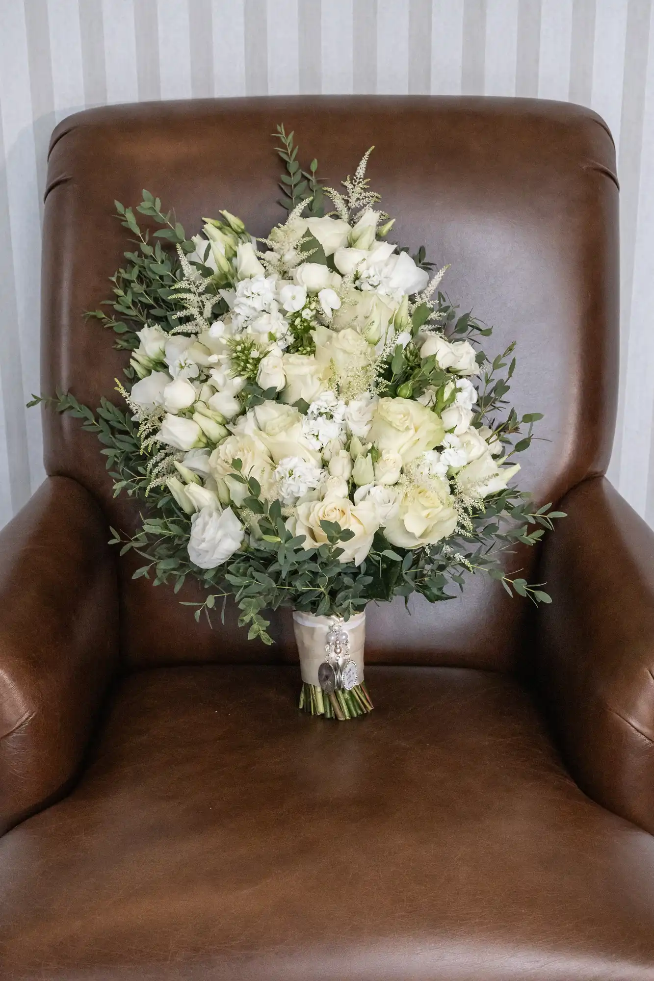 A bouquet of white roses and greenery sits on a brown leather chair against a striped wallpaper background.