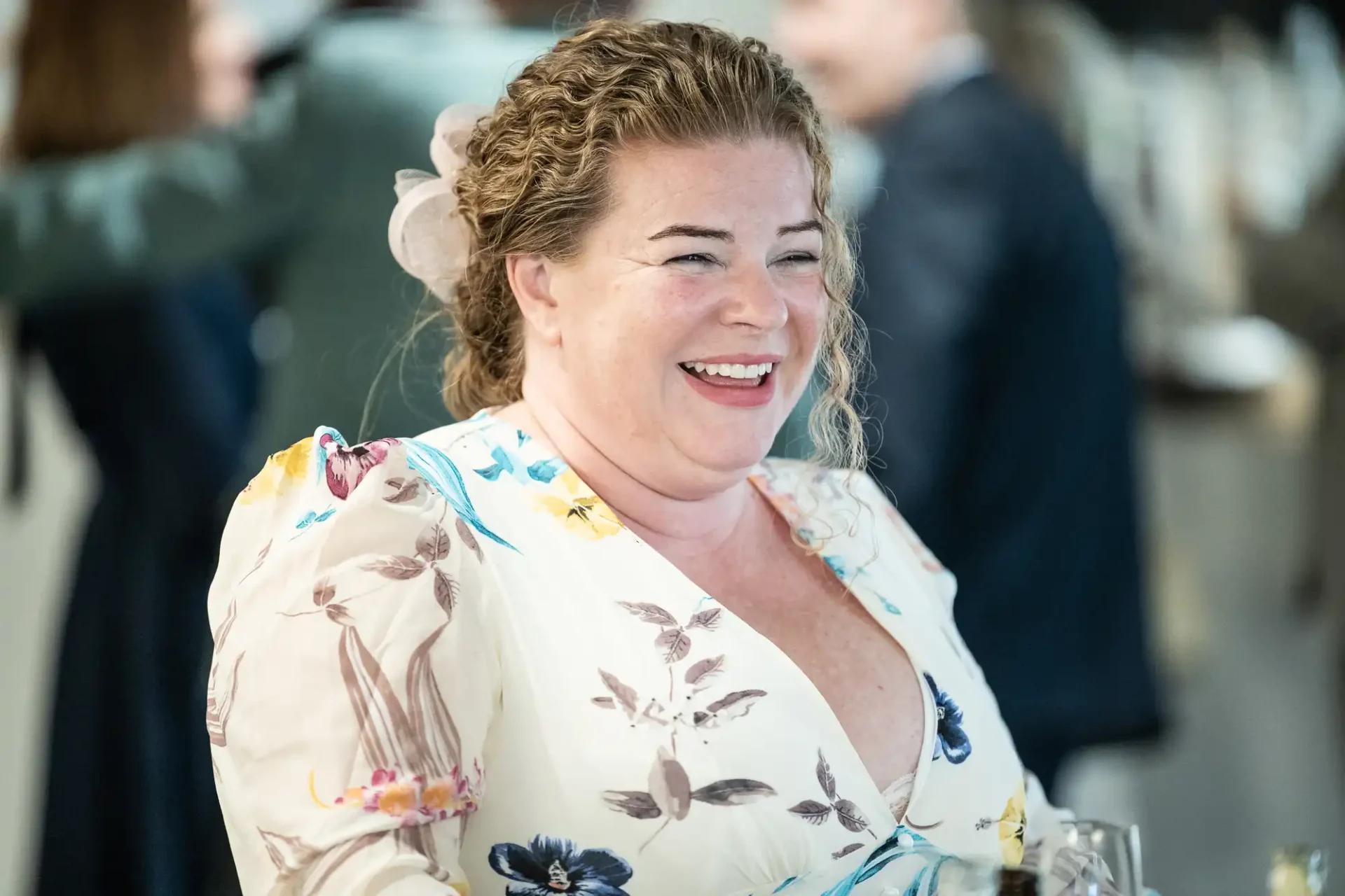 A woman with curly hair smiles, wearing a floral dress. She stands in a room with other people in the background.