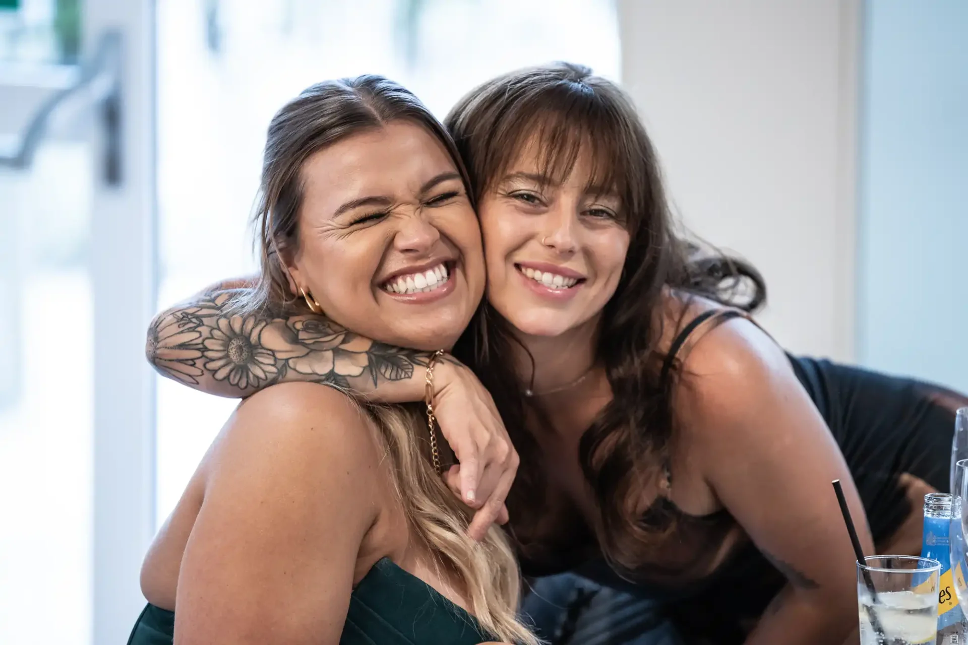 Two smiling women pose closely, one with a floral tattoo on her arm. Both appear happy, with drinks visible on the table in front of them.