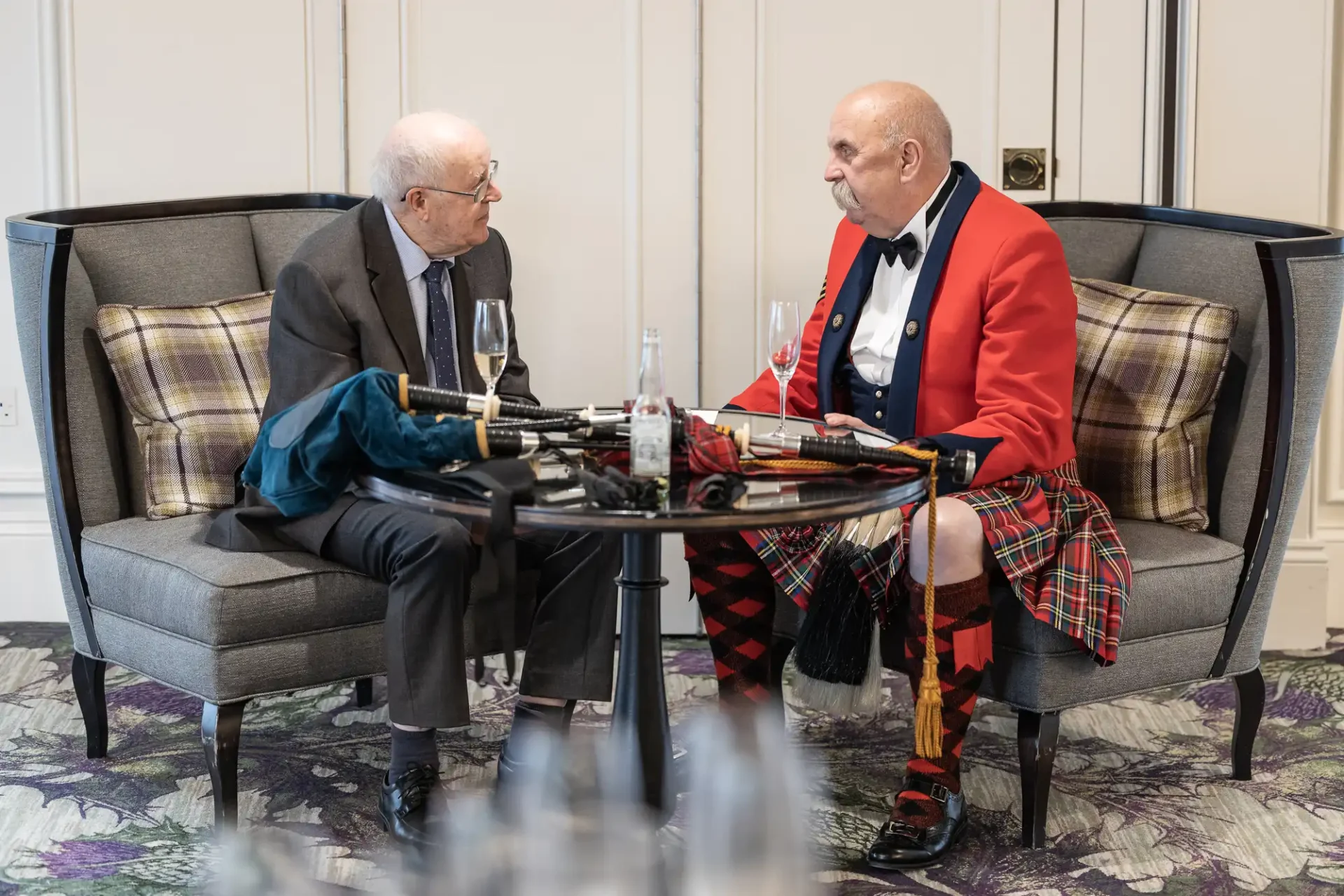 Two men sitting and conversing on a sofa. One wears a suit, the other a red military jacket with a kilt. Bagpipes rest on the table in front of them.