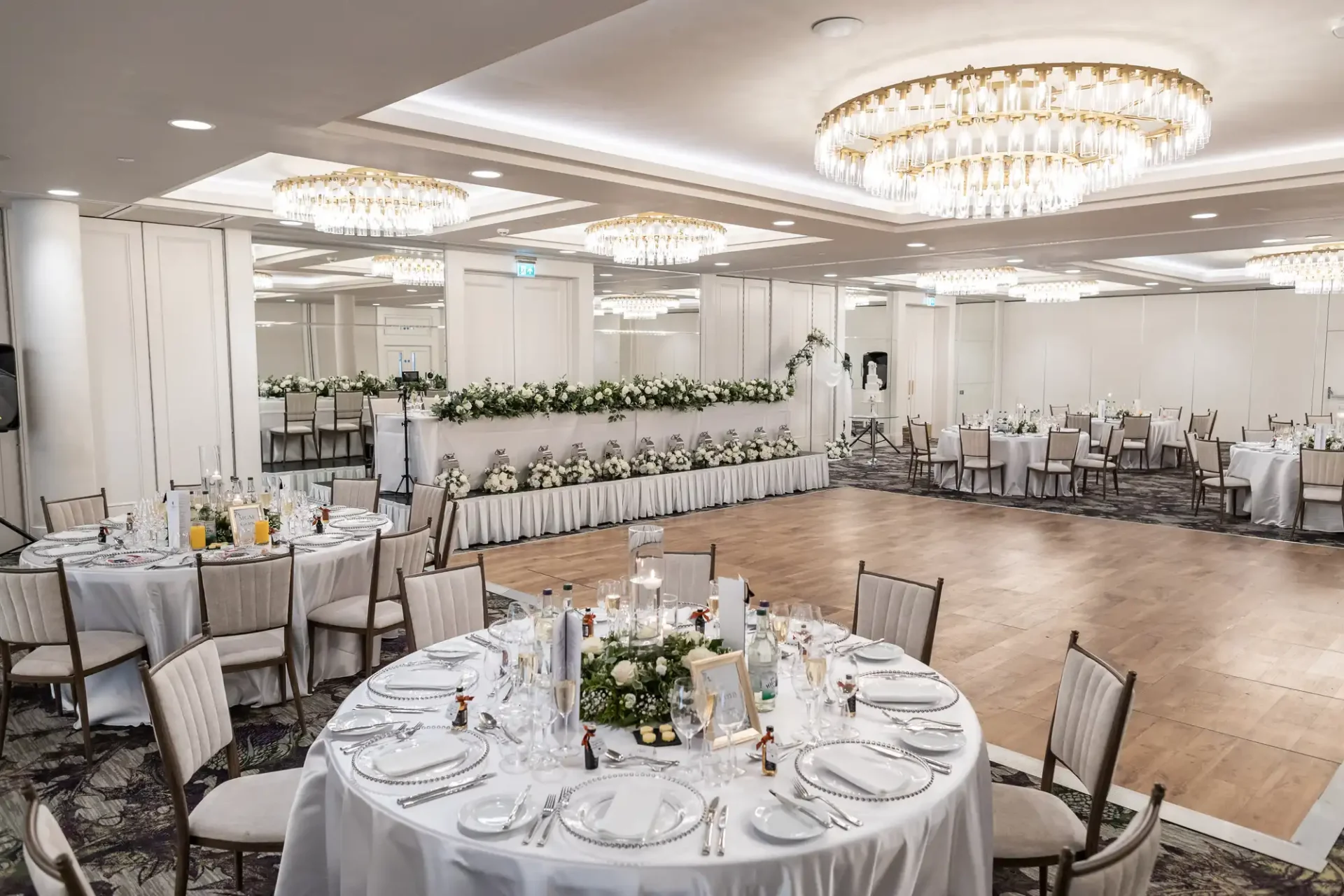 Elegant banquet hall with round tables set for an event, featuring white tablecloths, floral centerpieces, and chandeliers. Tables are arranged around a wooden dance floor.