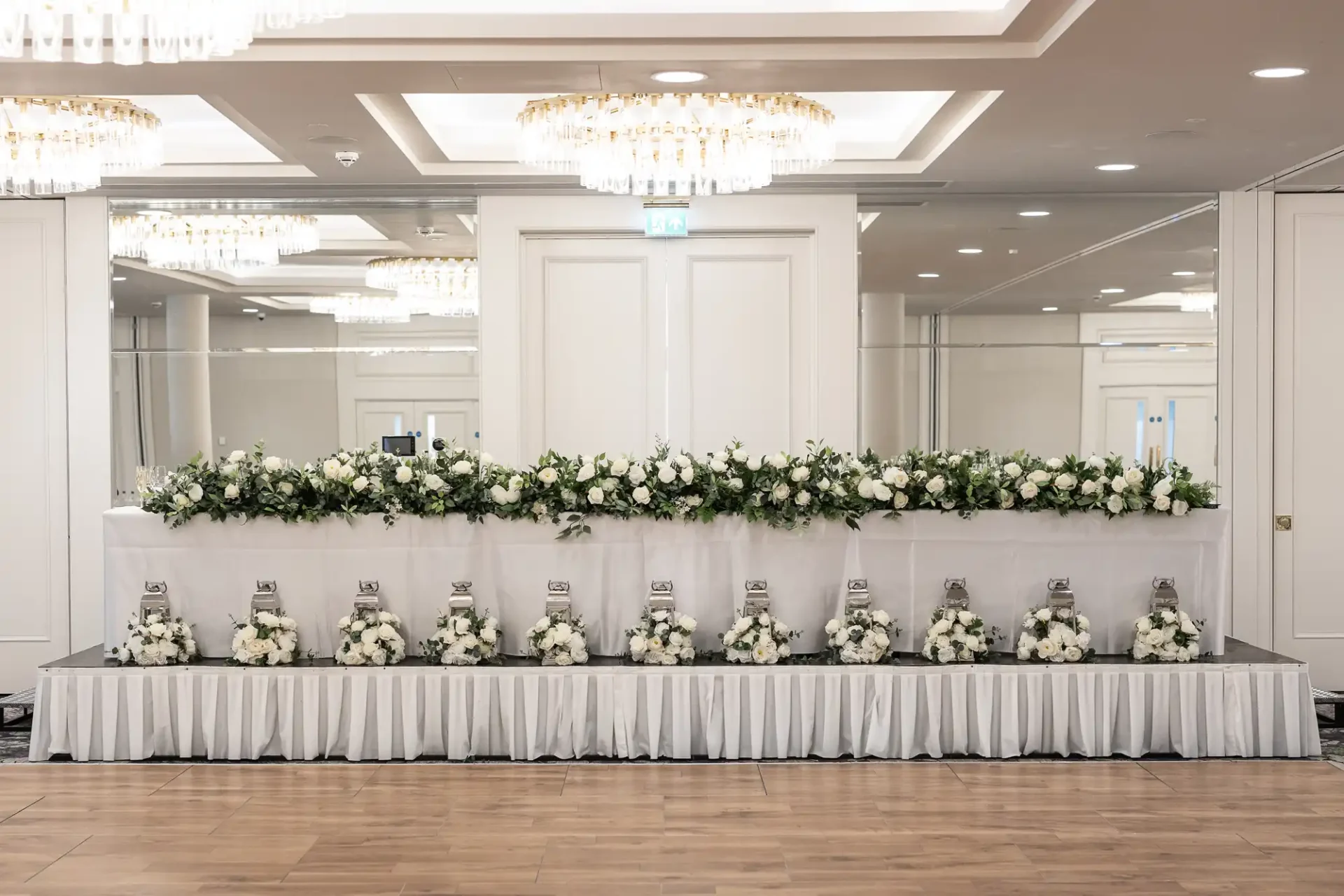 Elegant wedding reception table adorned with white flowers and greenery, beneath crystal chandeliers in a spacious, well-lit room with mirrored walls and wooden flooring.