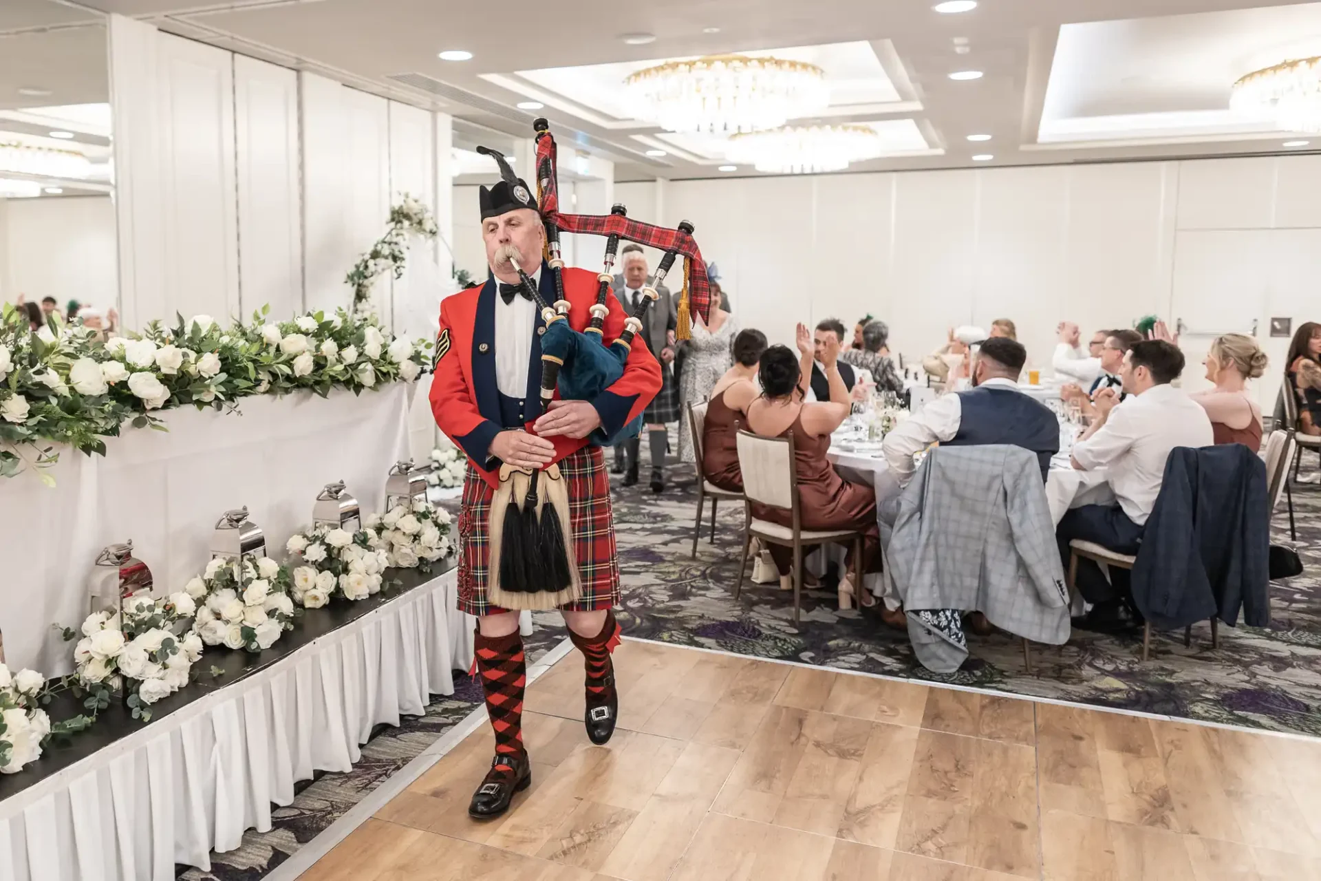 A bagpiper in traditional attire plays at a formal event, with seated guests attentively watching. Floral decorations line the room.