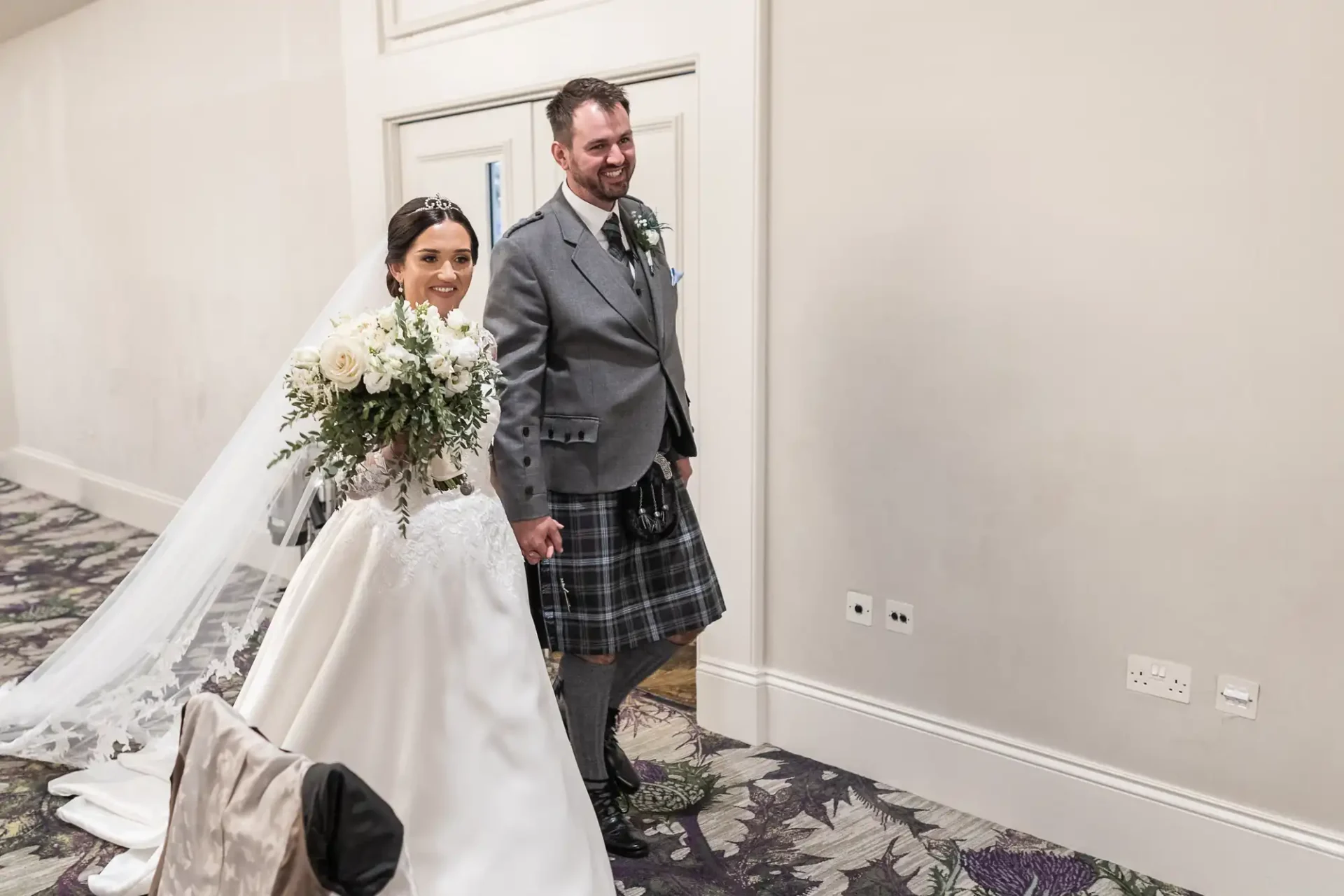 Bride in a white gown holding a bouquet and groom in a kilt walking indoors, holding hands, both smiling.