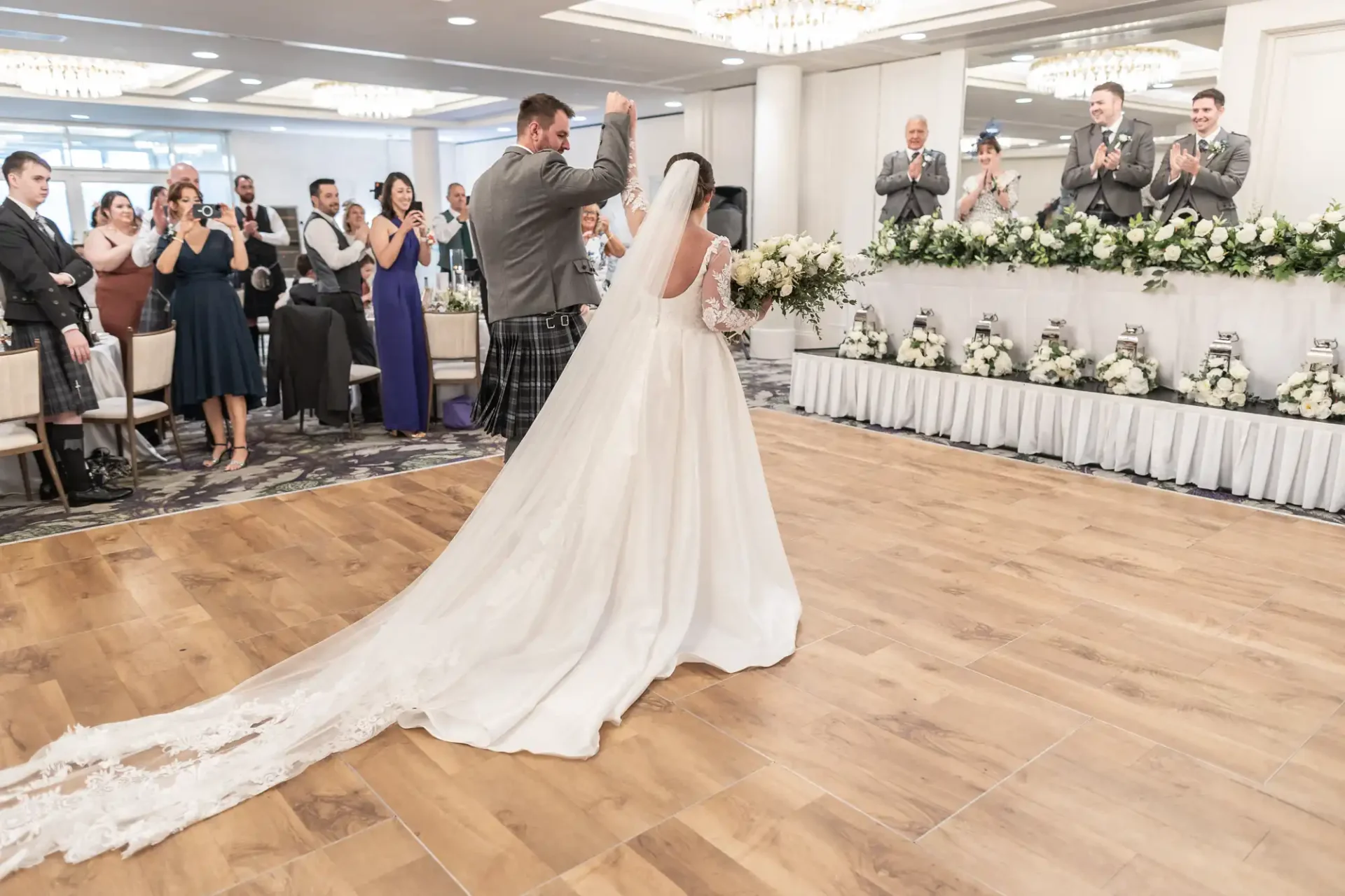 A bride and groom dance in a reception hall while guests and the bridal party applaud. The bride wears a long veil and dress; the groom is in a kilt. Floral arrangements decorate the room.