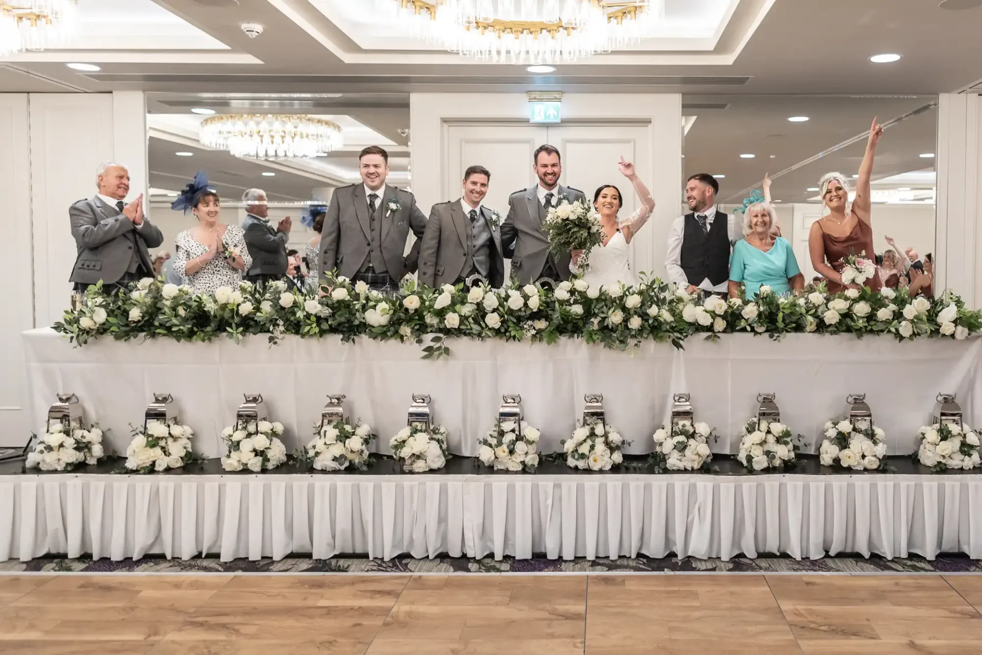 A wedding party standing behind a long table adorned with white floral arrangements. All are dressed in formal attire, and some are raising their hands in celebration.