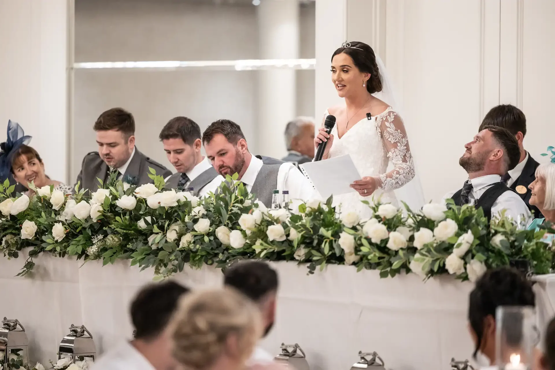 A bride in a white dress gives a speech at a decorated table. Several people sit beside her, with white roses and greenery in front.