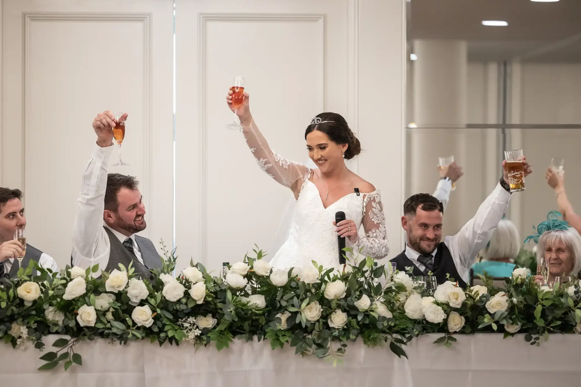 Bride in a white dress holds a microphone and raises a glass, surrounded by smiling guests holding drinks, at a decorated table with white flowers.