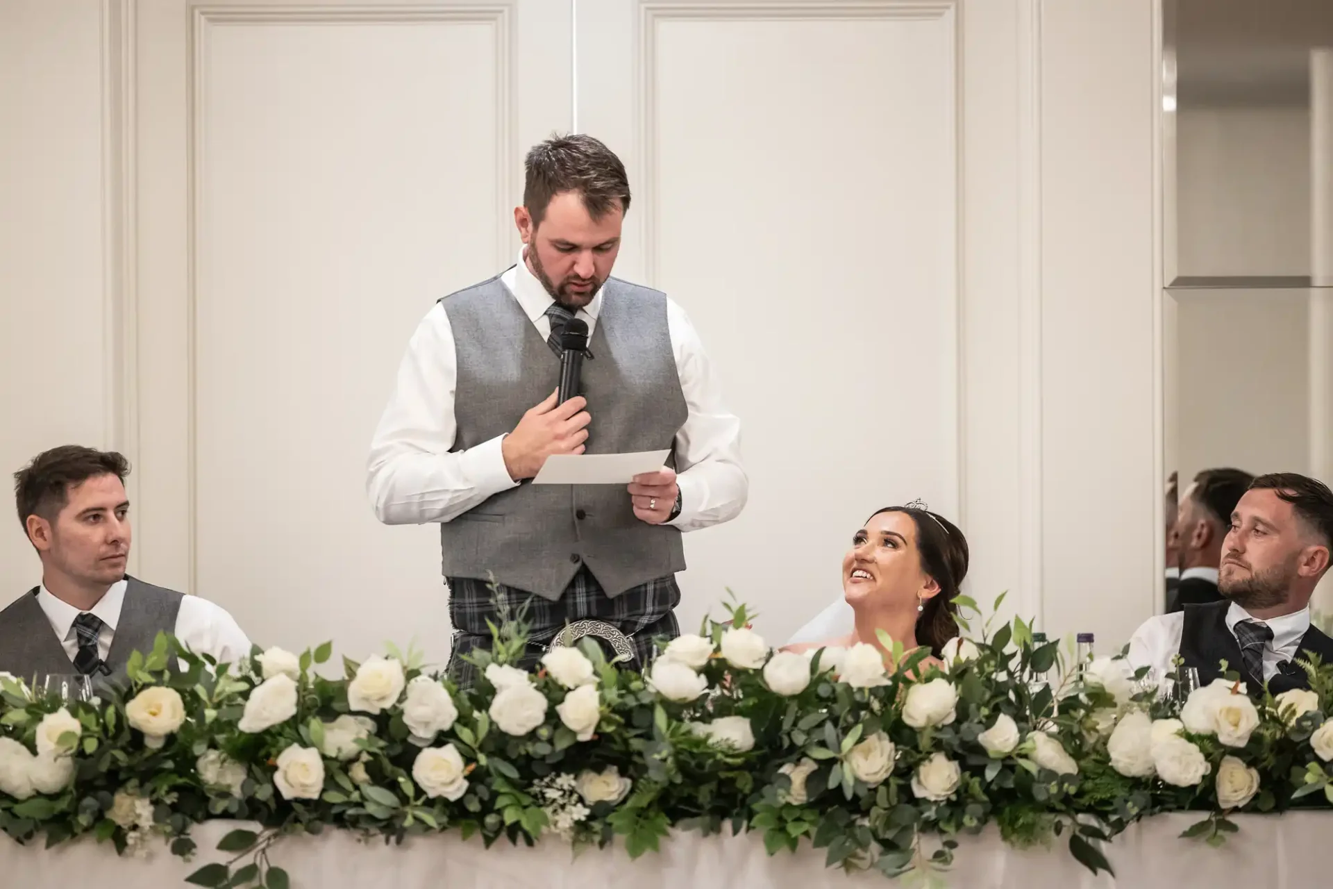 A man in a suit vest gives a speech at a wedding reception. He holds a microphone and paper. Three people sit at a table decorated with white flowers and greenery, listening to him.