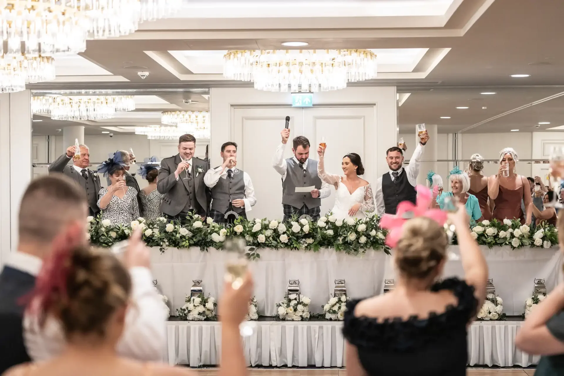 A wedding party raises glasses in a toast. The bride and groom are at the center, surrounded by the bridal party at a decorated table with white flowers. Guests are visible in the foreground.