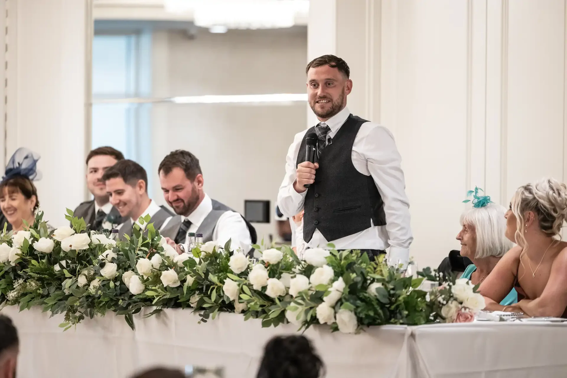 A man in a vest and tie gives a speech at a wedding reception. He stands behind a table adorned with white flowers. Several people are seated beside him, listening.