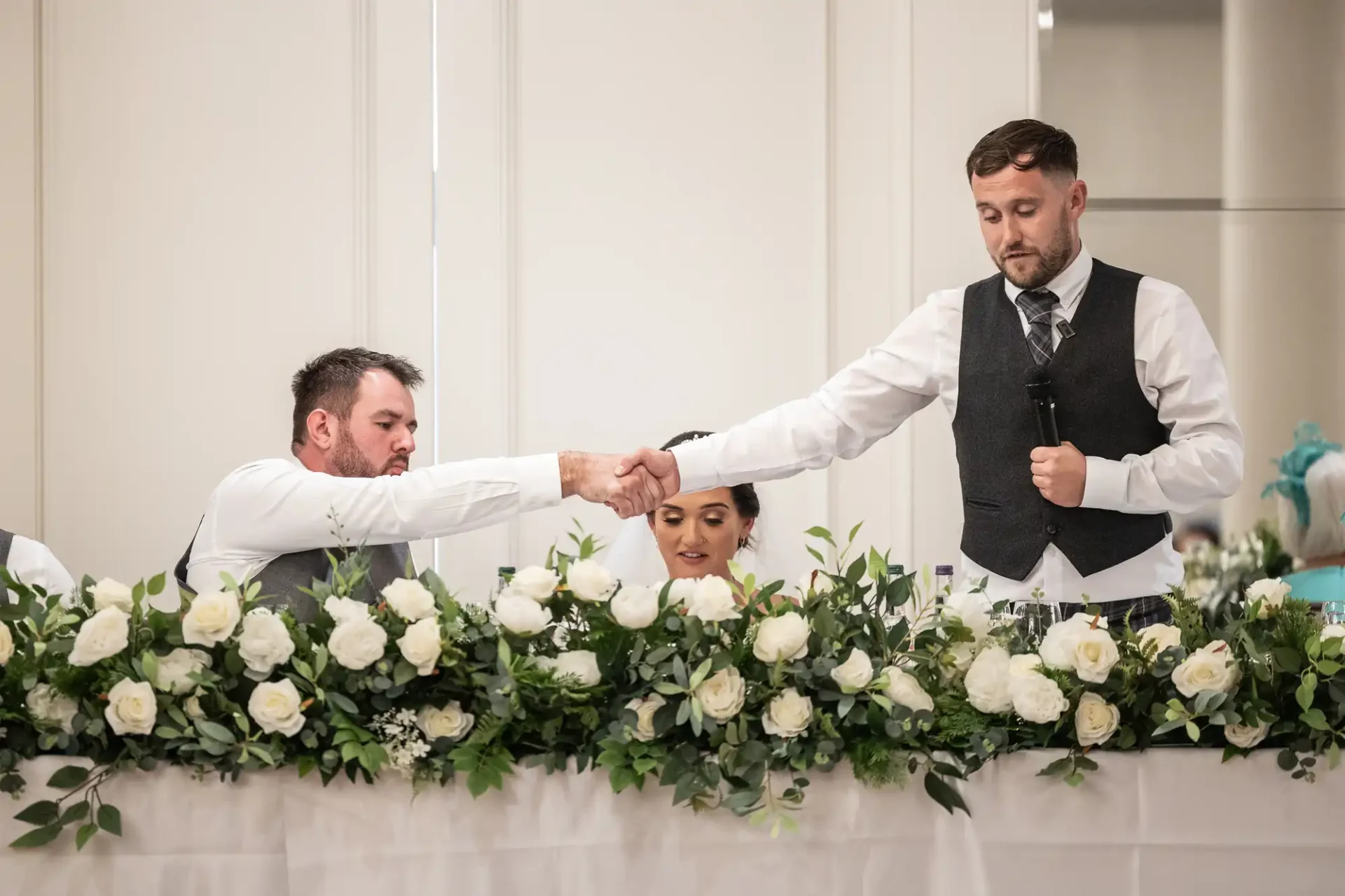 A man in a vest gives a speech with a microphone, shaking hands with another man across a table adorned with white flowers. A woman sits in between them, looking down.