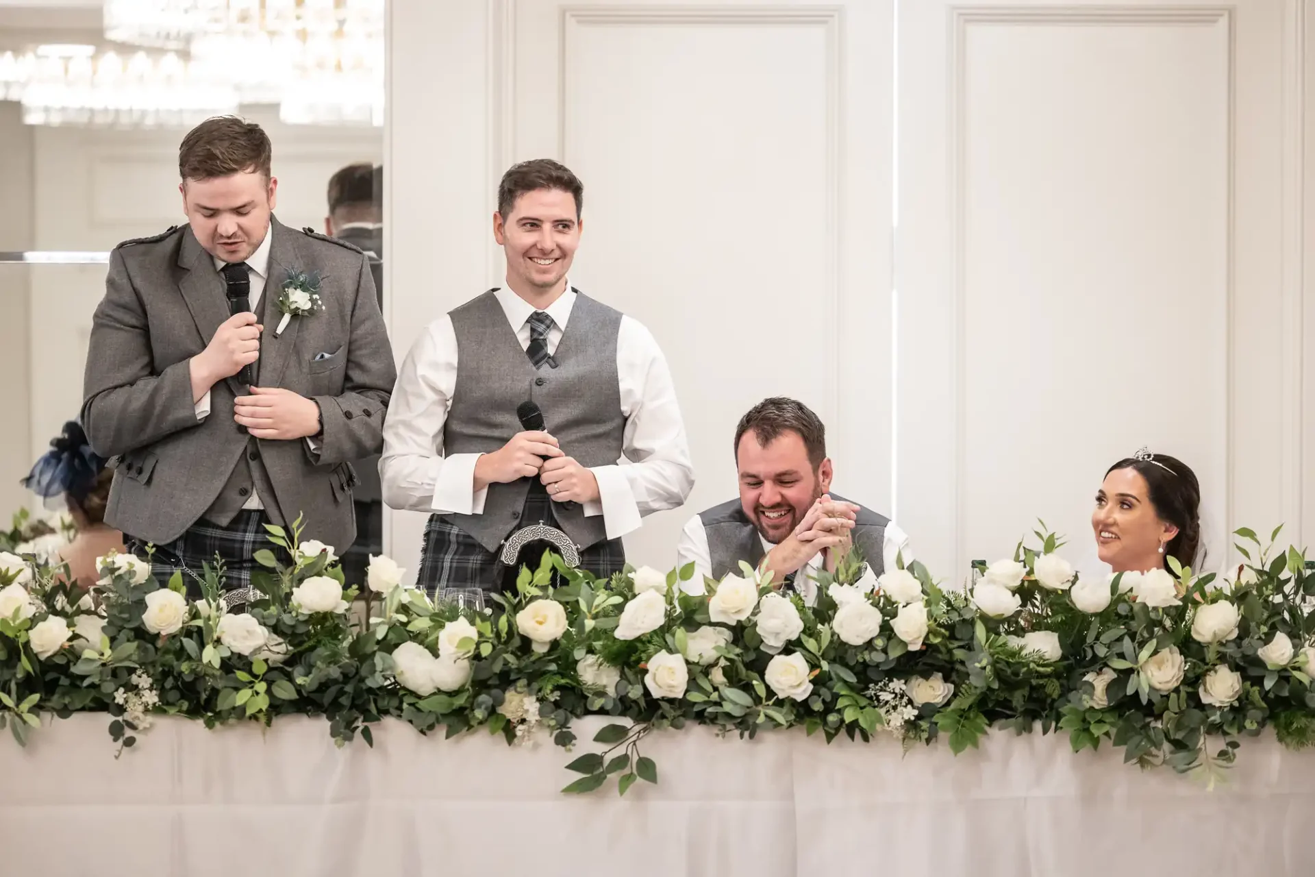 Four people seated at a table with floral decorations, two men are standing and speaking into microphones, while the other man and woman seated look on.