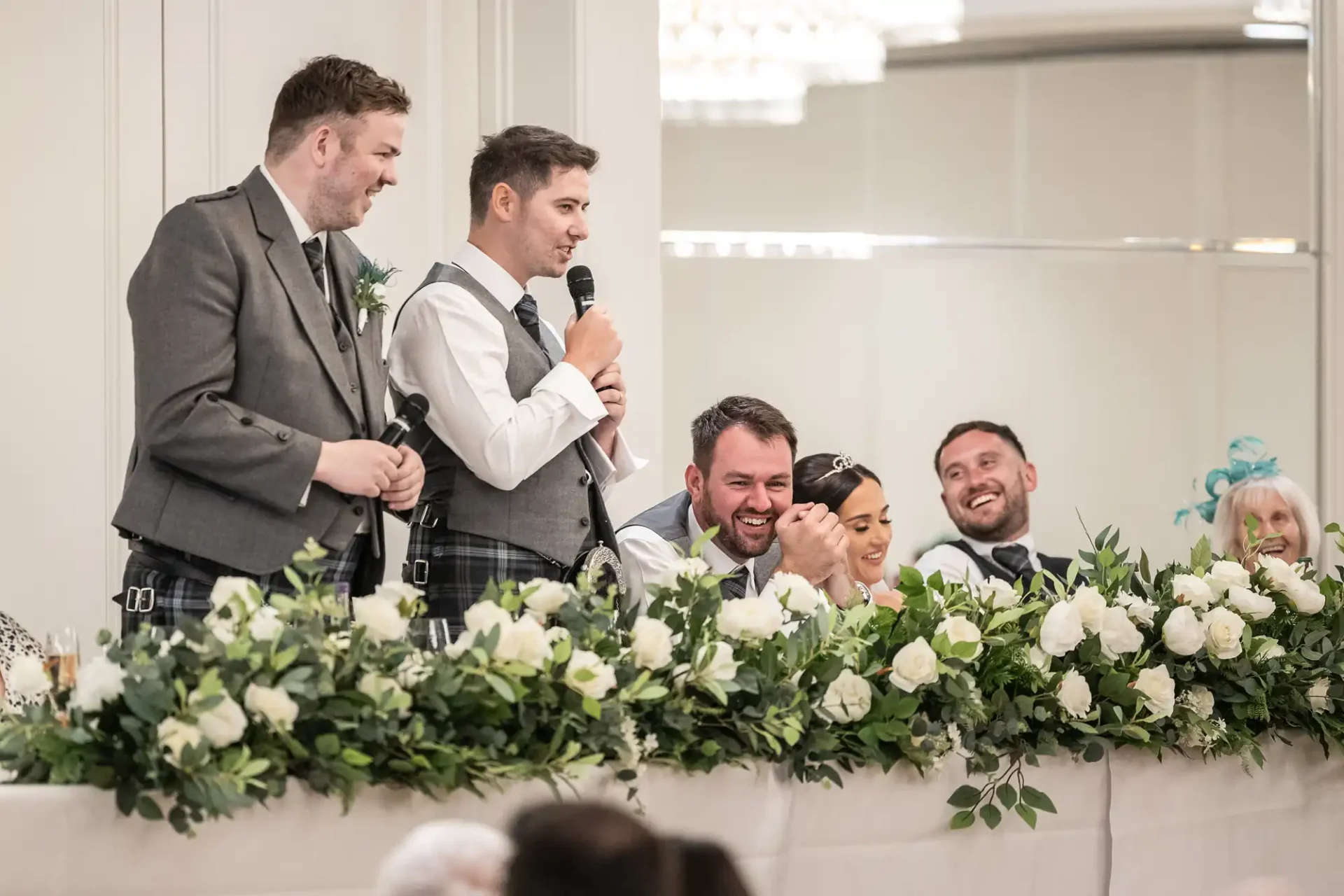 People in formal attire are seated at a decorated table during a wedding reception. A man standing with a microphone is speaking, while others listen and smile.