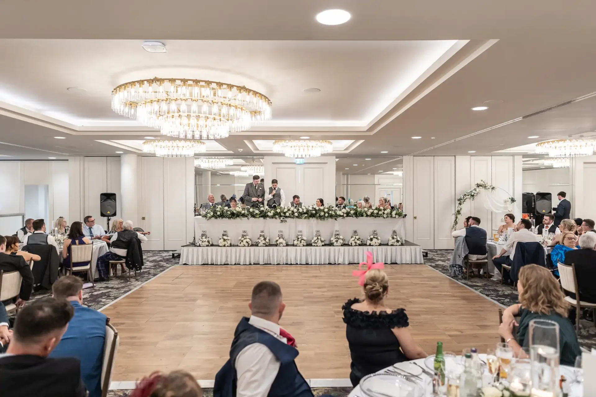 A wedding reception in a decorated banquet hall with guests seated around tables. A long table with floral arrangements is at the front, with speakers addressing the audience.