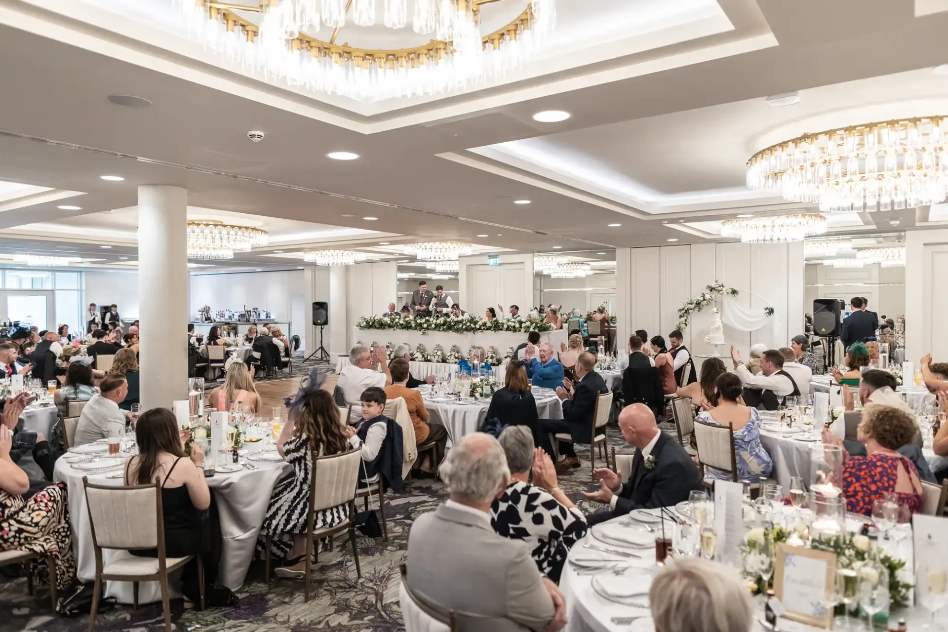 Large banquet hall filled with seated guests at decorated tables, watching a wedding reception speech. Chandeliers hang from the ceiling, and floral arrangements adorn the head table.