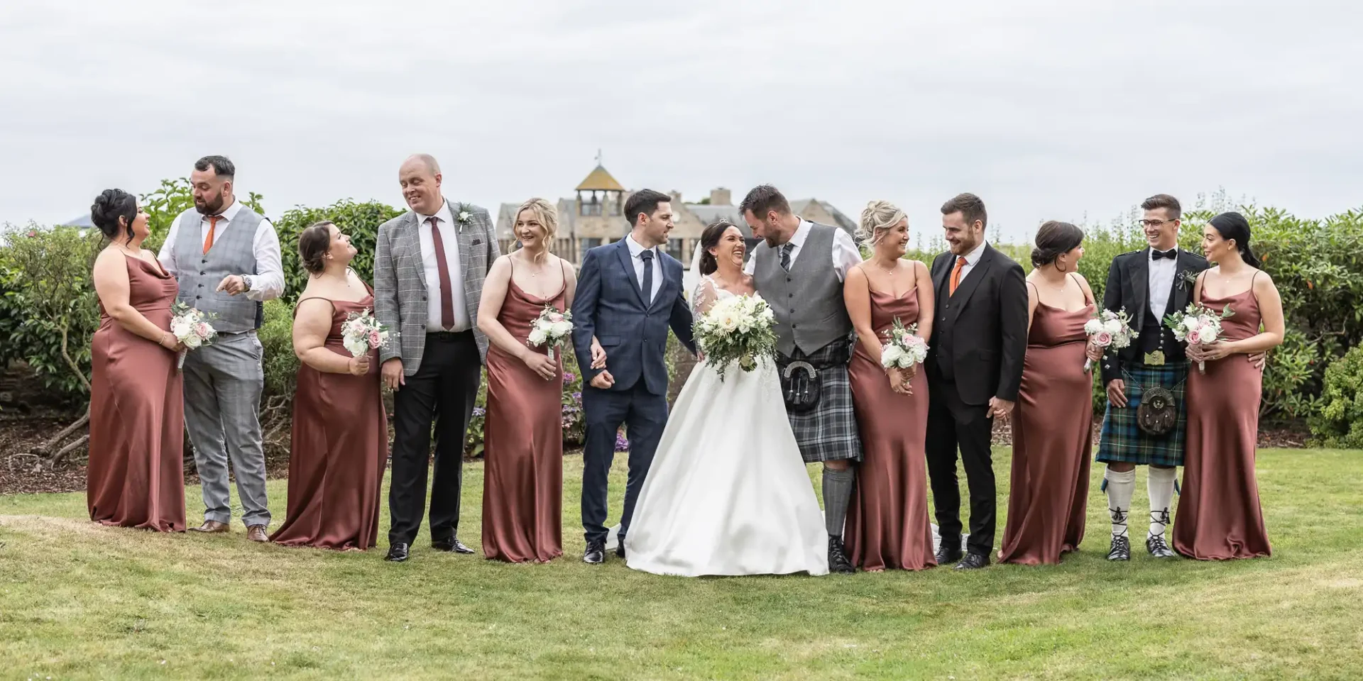 Bridal party stands in a line outdoors, with bridesmaids in brown dresses and groomsmen in various suits, including kilts. The bride and groom are in the center, smiling at each other.