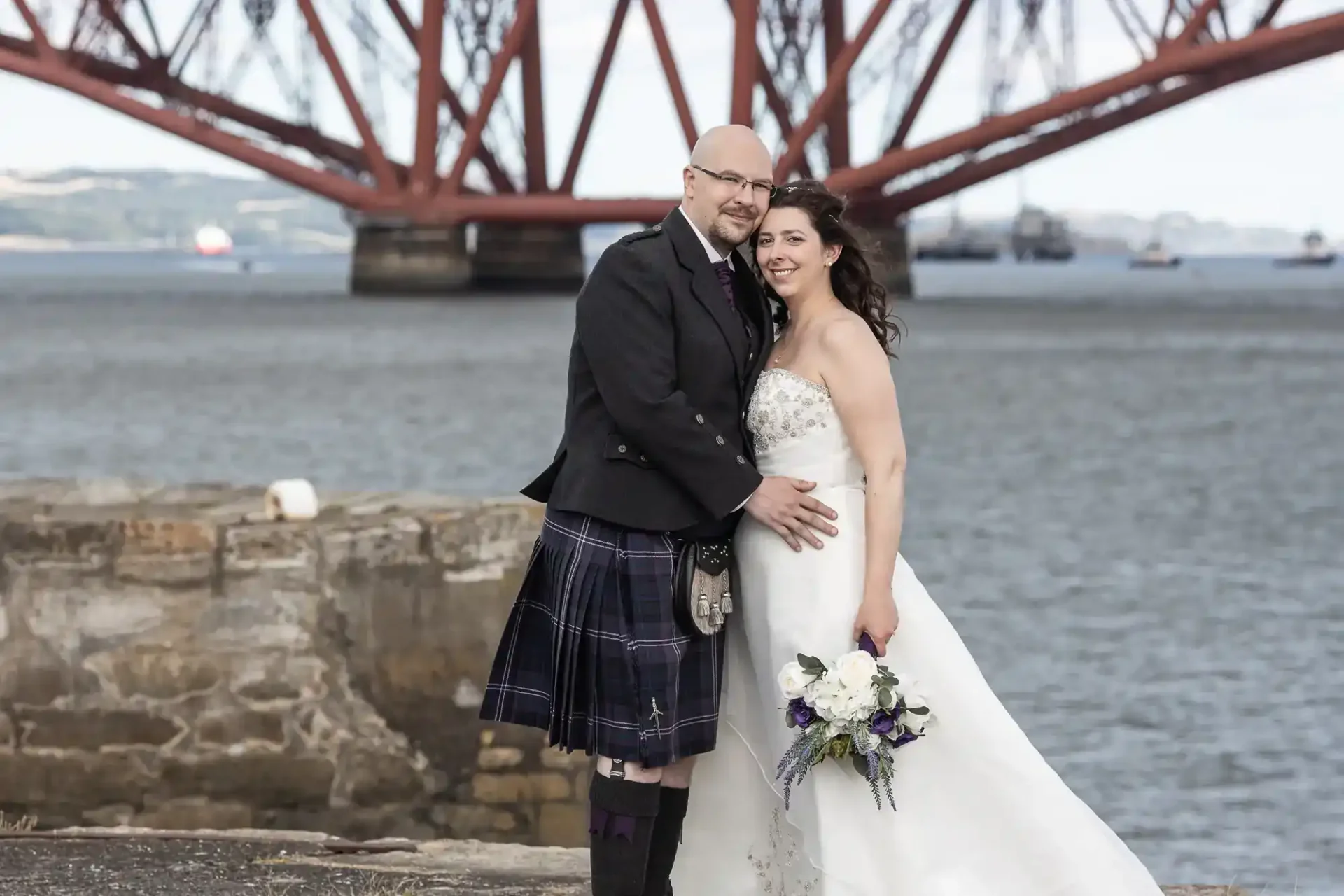 A couple in wedding attire poses by the water, with a large red bridge in the background. The groom wears a kilt; the bride holds a bouquet.