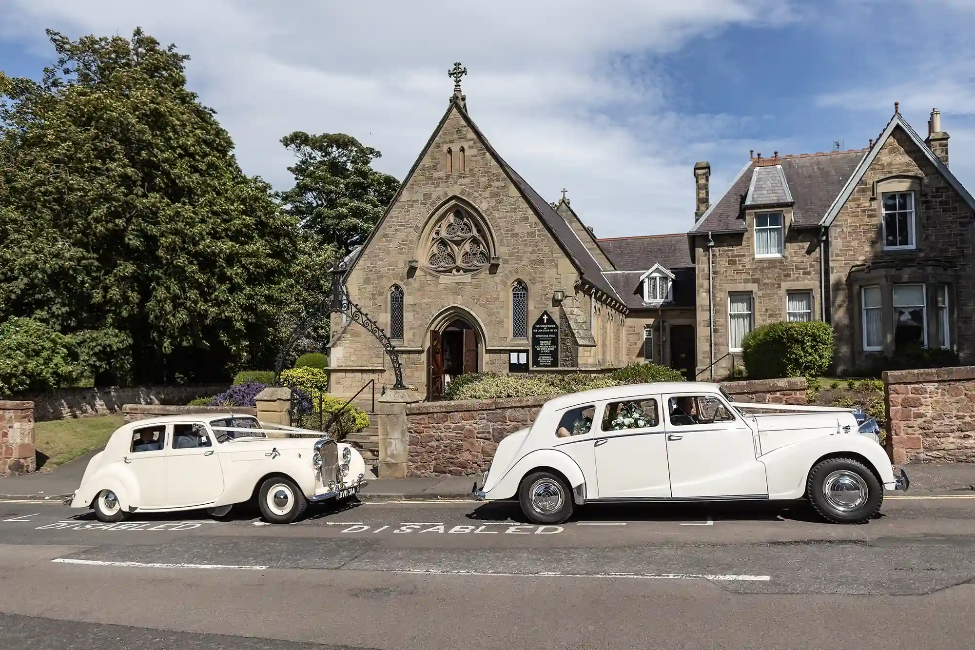 Two vintage white cars parked outside a quaint stone church with a sign marked "disabled" on the road, under a clear blue sky.