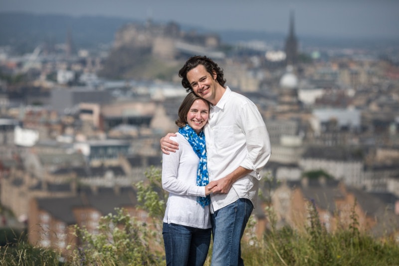 A couple embracing and smiling at the camera with edinburgh's cityscape, including the castle, in the background.
