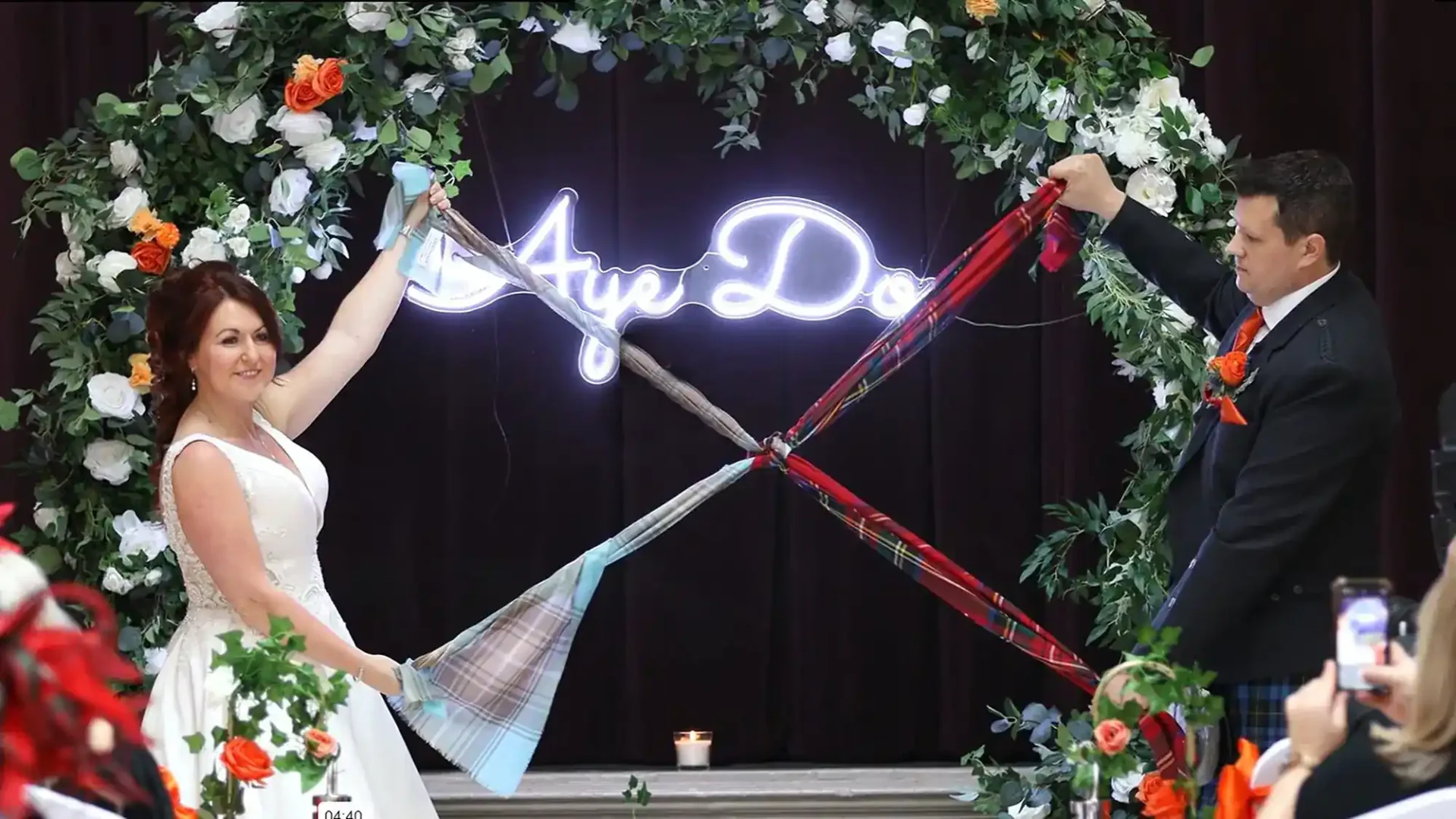 Peebles Hydro wedding video: Bride and groom hold tartan ribbons, standing under a floral arch with a neon "Aye Do" sign.