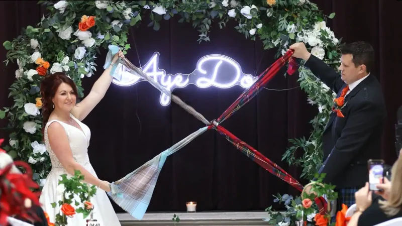 Bride and groom hold tartan ribbons, standing under a floral arch with a neon "Aye Do" sign.