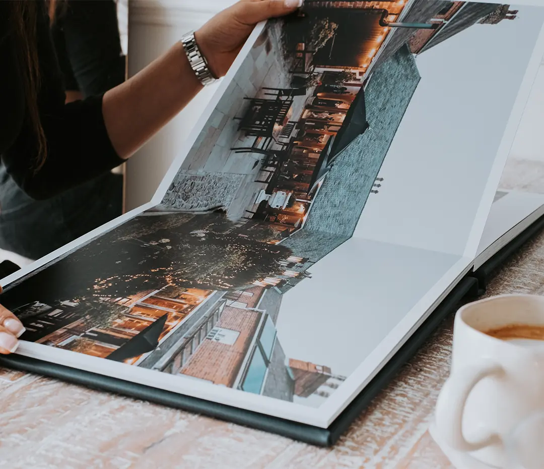 A person with a watch is holding open a photo album displaying two cityscape images, next to a white coffee mug on a wooden table.