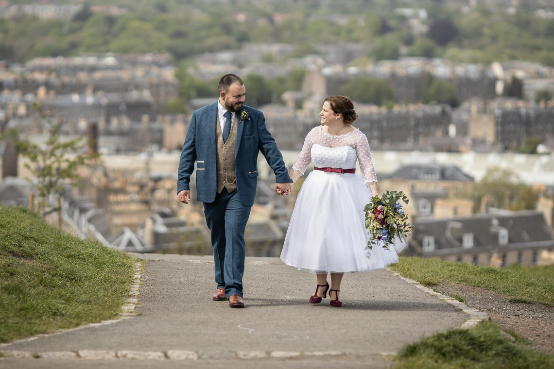 Photographer in Edinburgh Calton Hill Sarah and Tom - a bride and groom walking hand in hand on a pathway overlooking a cityscape, the bride holding a bouquet, both dressed in wedding attire.