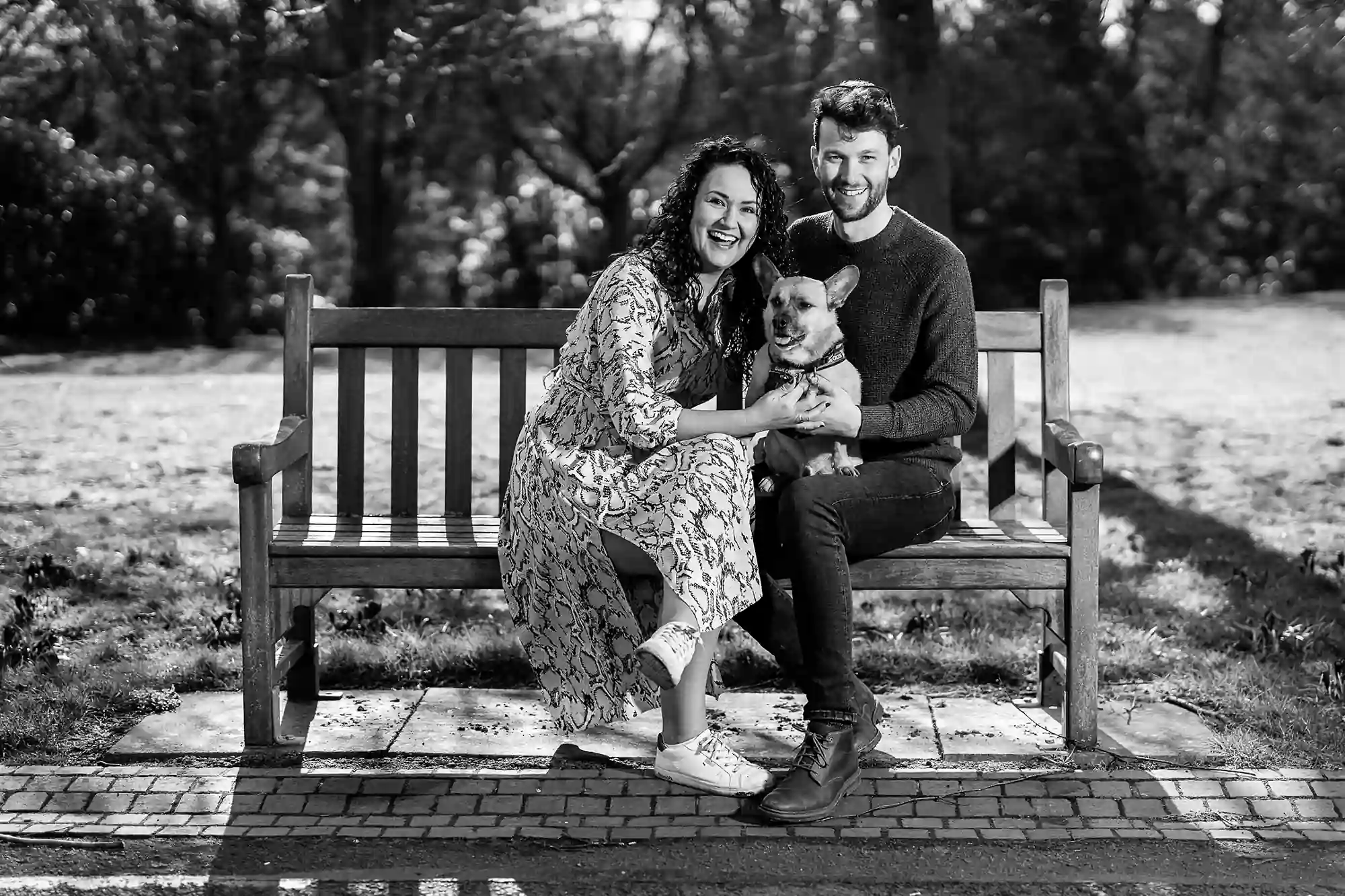 A happy couple and their dog sitting on a park bench, smiling towards the camera in a sunny, tree-lined setting.