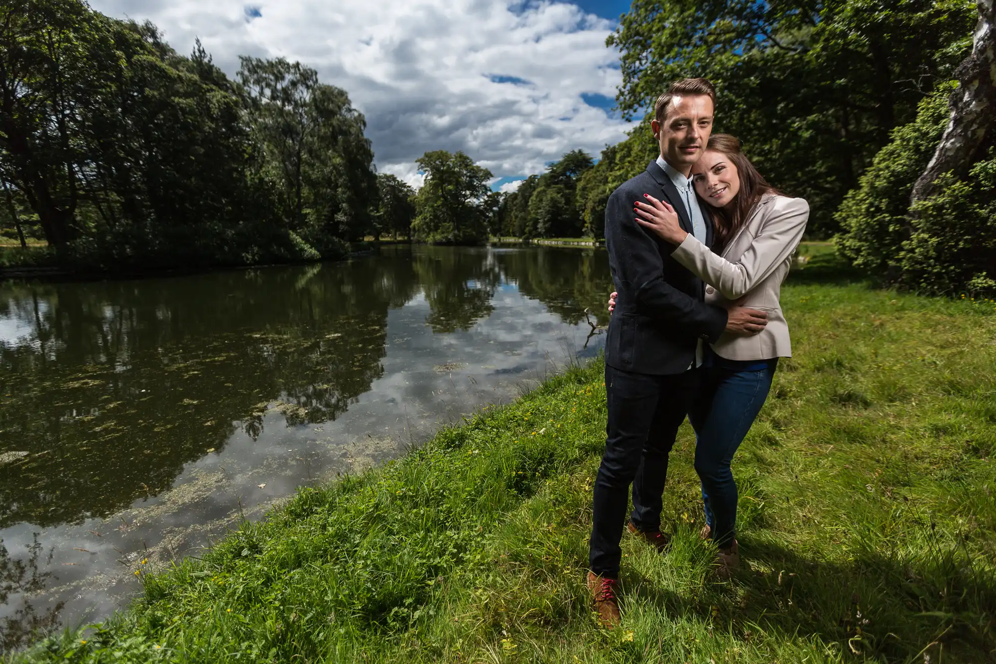 A couple embracing near a serene river surrounded by lush greenery under a cloudy sky.