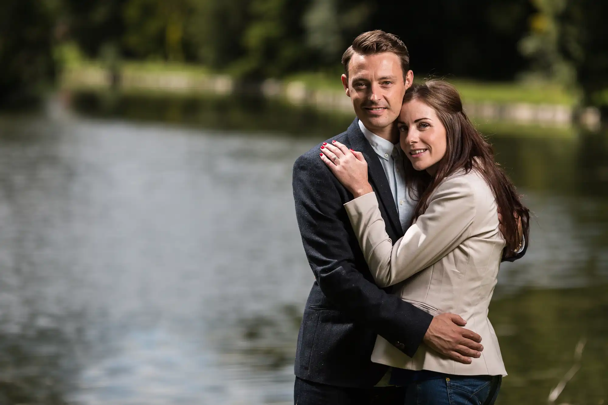 A couple embracing affectionately by a tranquil lake. the man, in a suit, smiles at the camera while the woman, in a beige jacket, rests her head on his chest.
