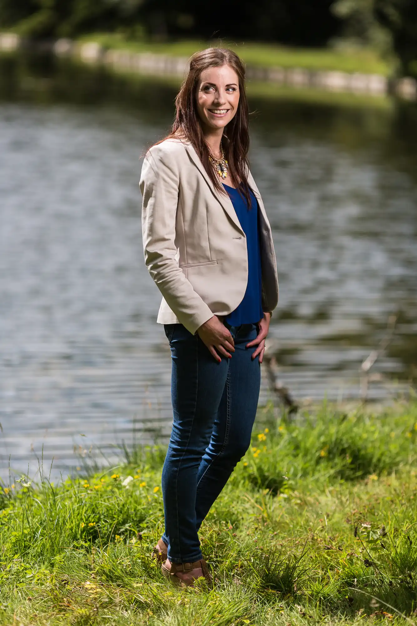 A woman in a blazer and jeans smiling by a lakeside.