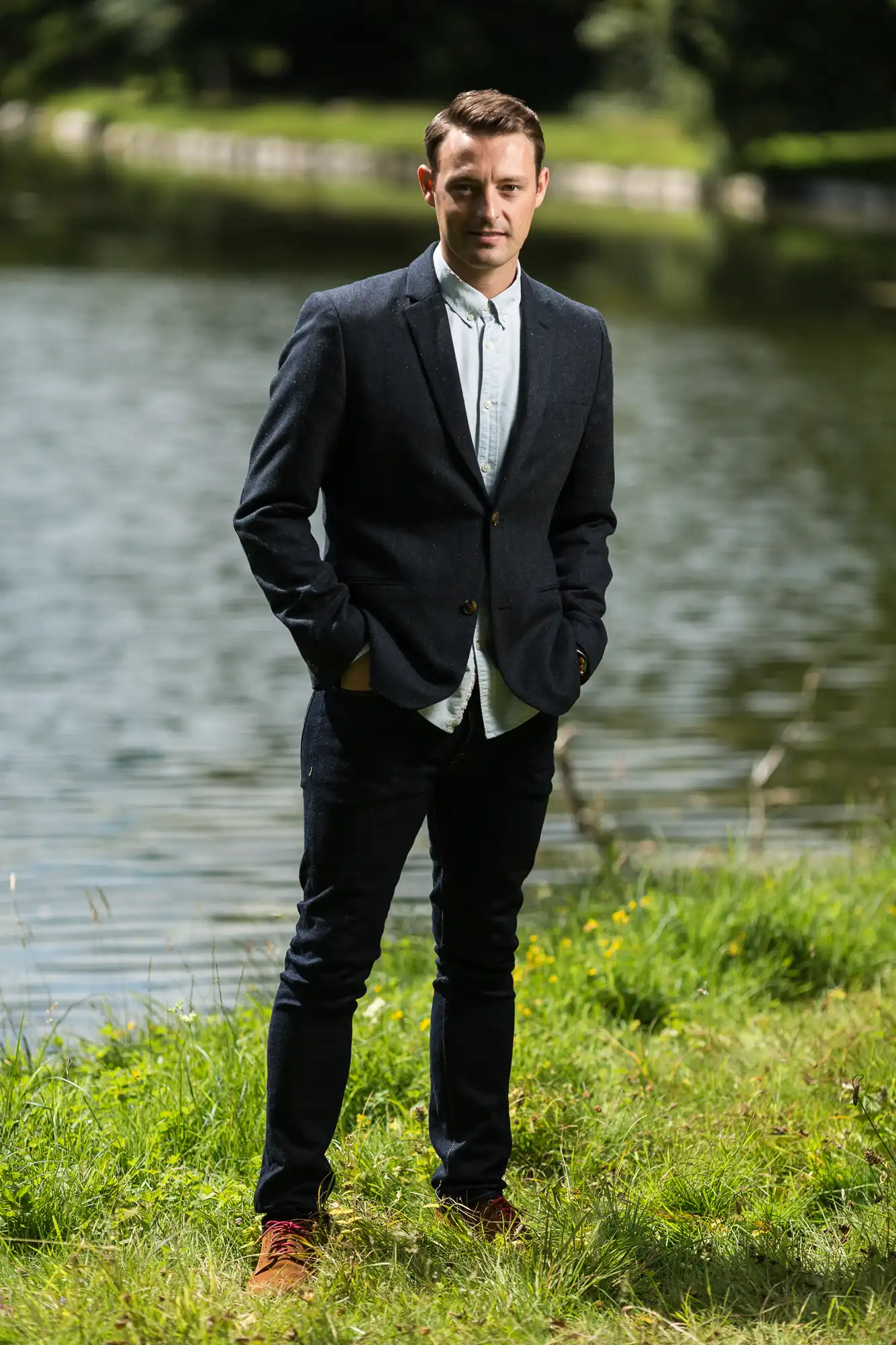 A man in a smart casual outfit stands confidently by a pond, hands in pockets, surrounded by lush greenery.