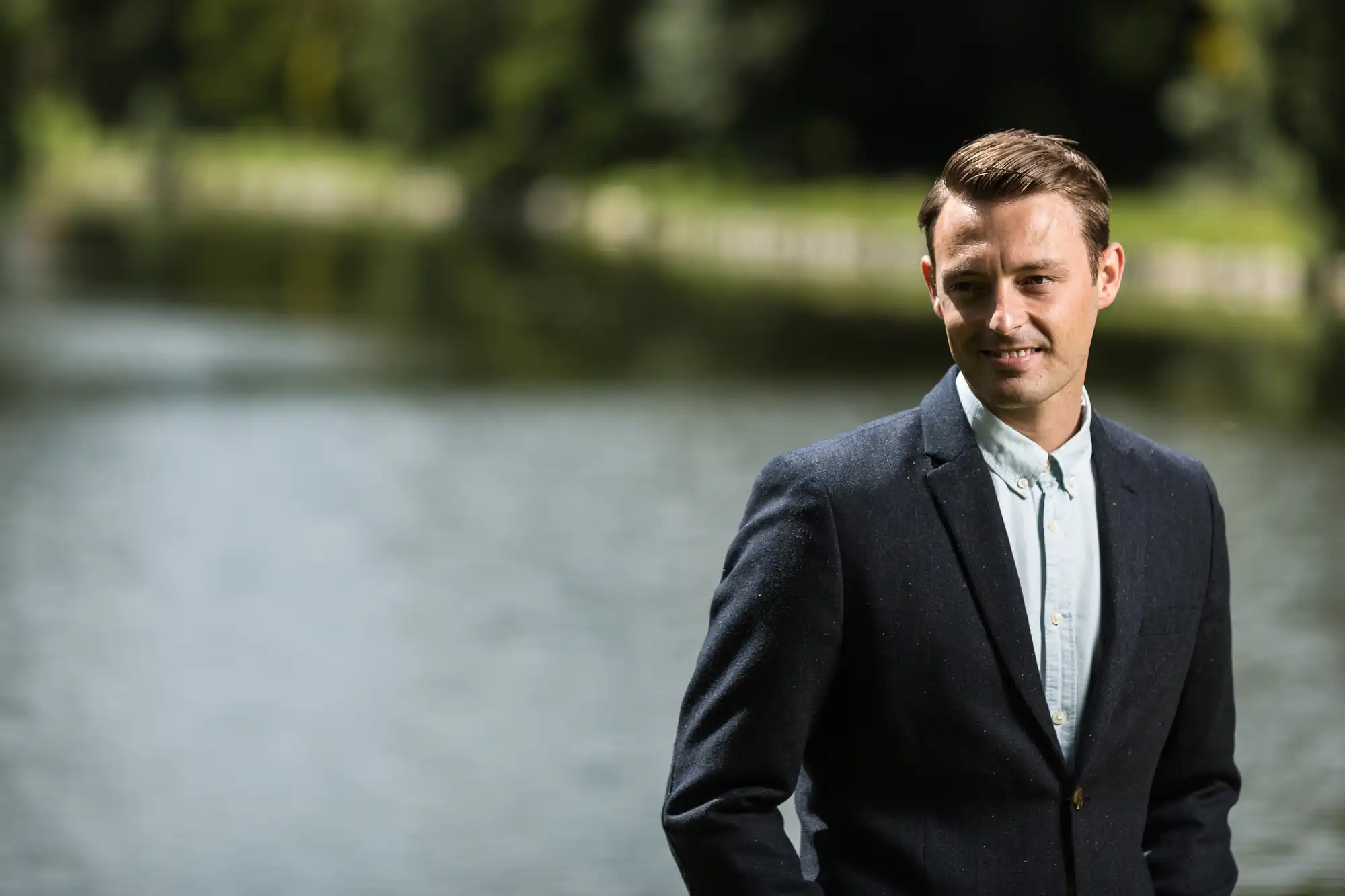 A young man in a smart blazer and button-up shirt, smiling gently by a serene river backdrop.