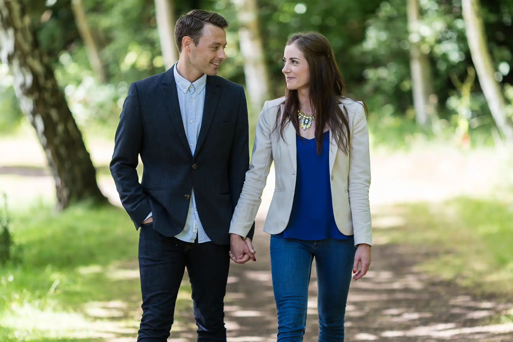 A man in a suit and a woman in a blazer and jeans walking hand in hand on a wooded path, smiling at each other.