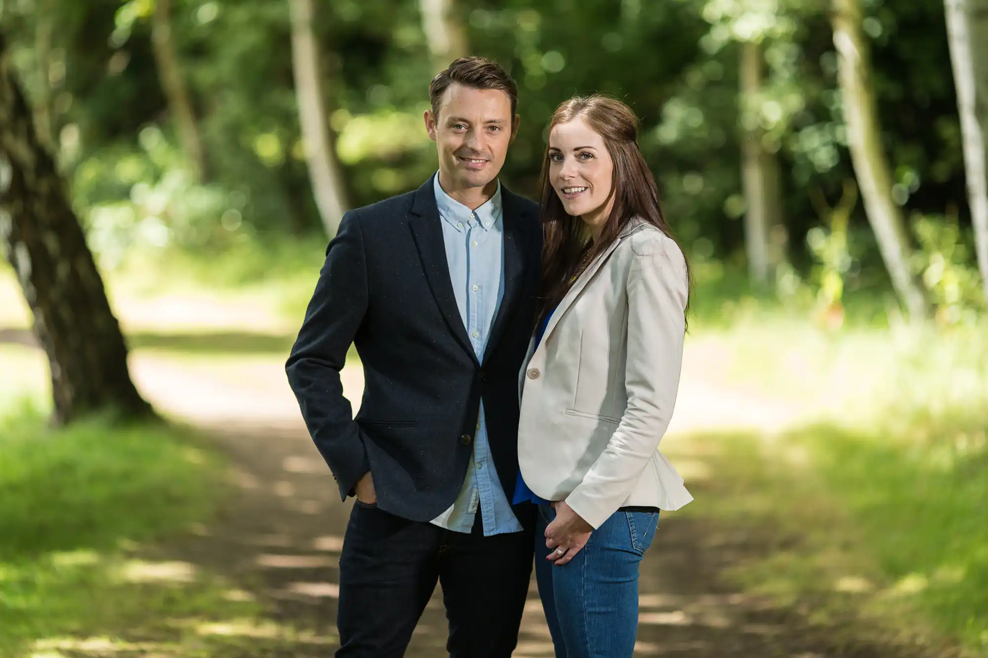 A man and a woman standing together on a forest path, both smiling at the camera. the man wears a blue blazer, and the woman is in a white jacket and jeans.