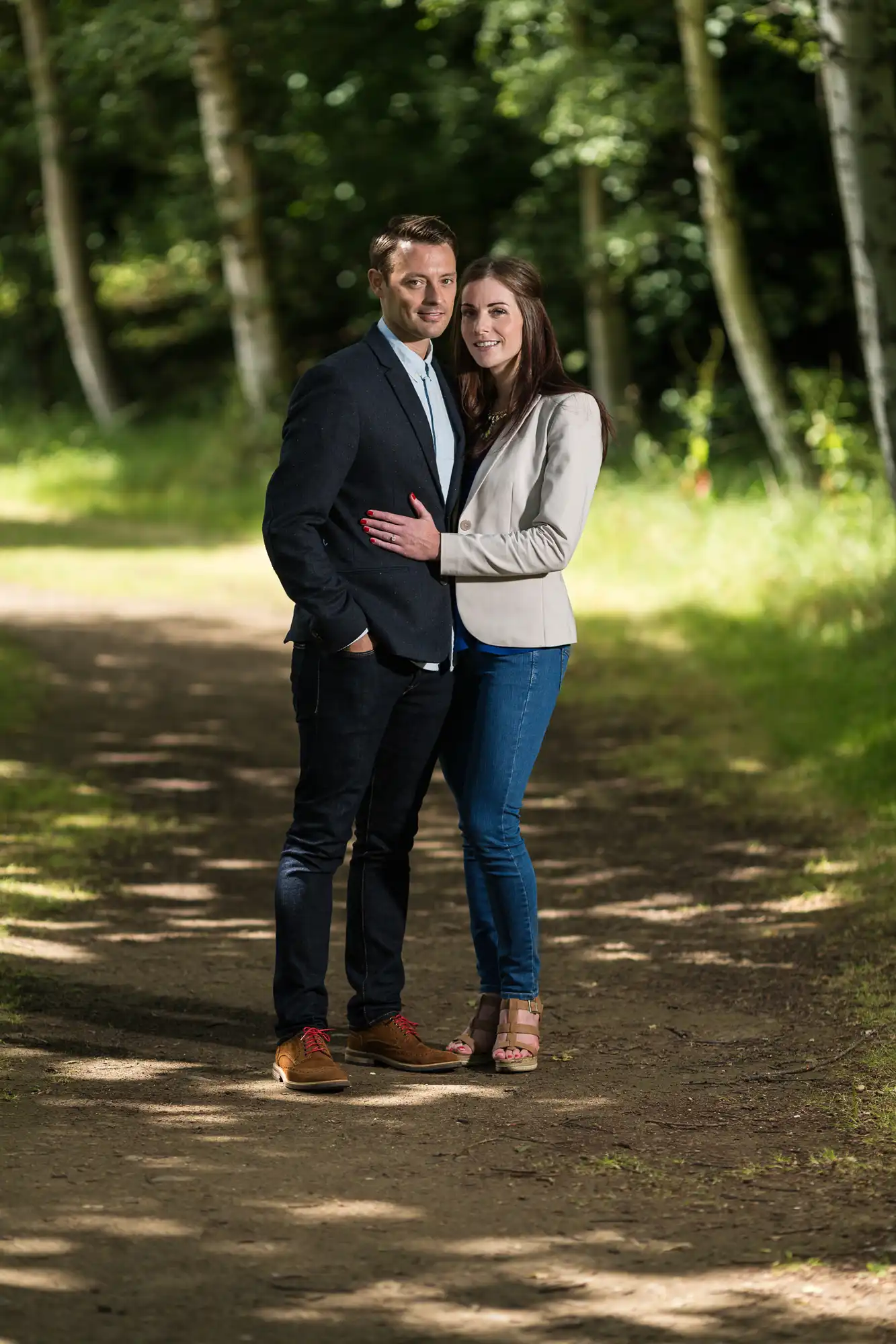 A couple embracing on a forest path, with the man in a blazer and jeans, and the woman in a blazer and blue jeans, both smiling at the camera.