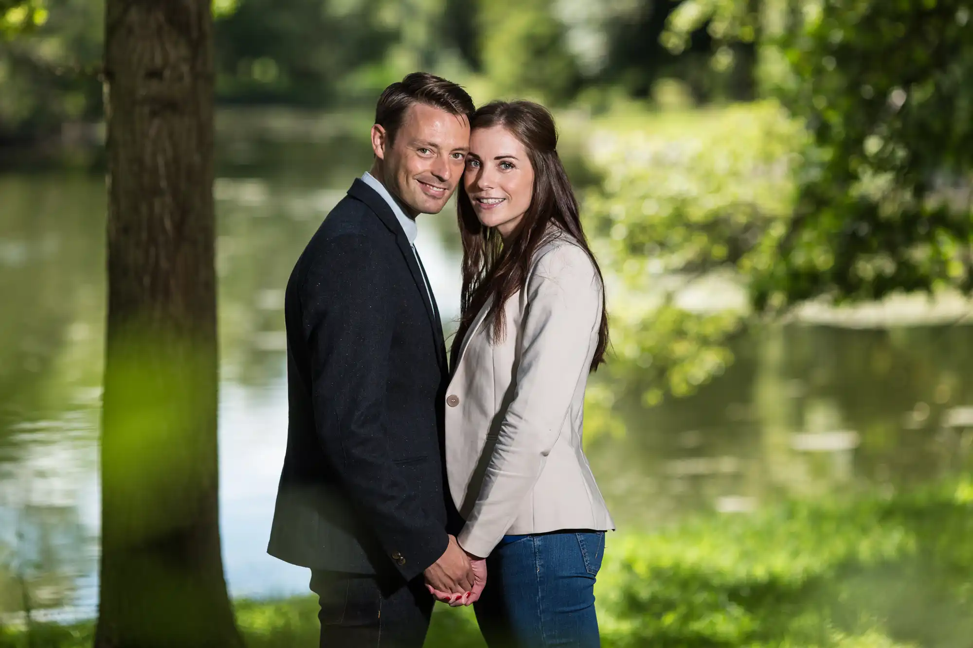 A couple holding hands and smiling at the camera, standing beside a serene lake surrounded by green trees.