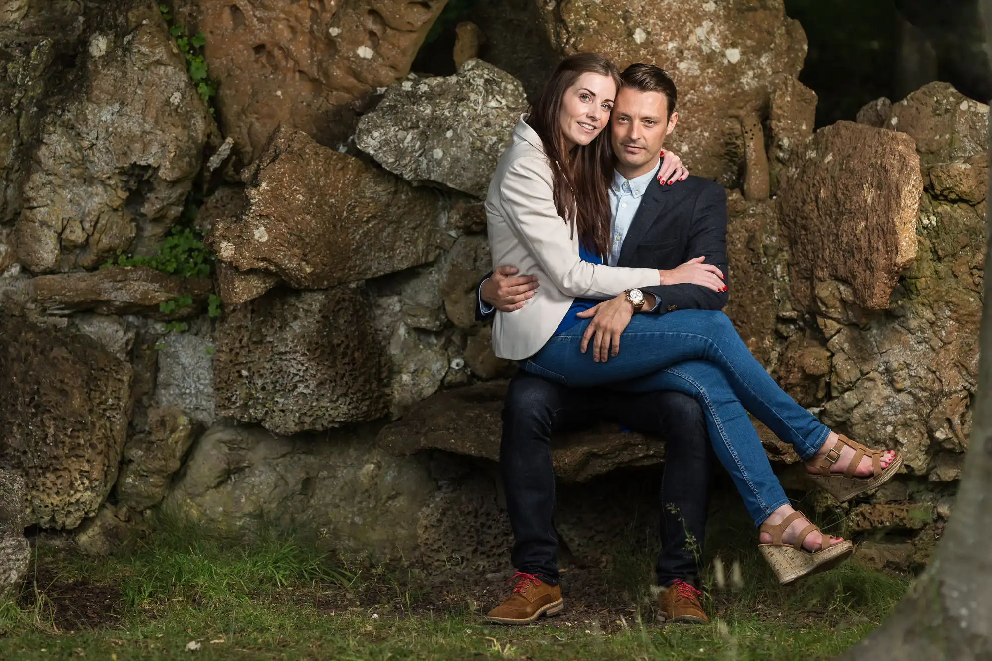 A couple sitting closely together, the man seated on a rock and the woman embracing him from behind, both smiling, set against a rugged stone backdrop.