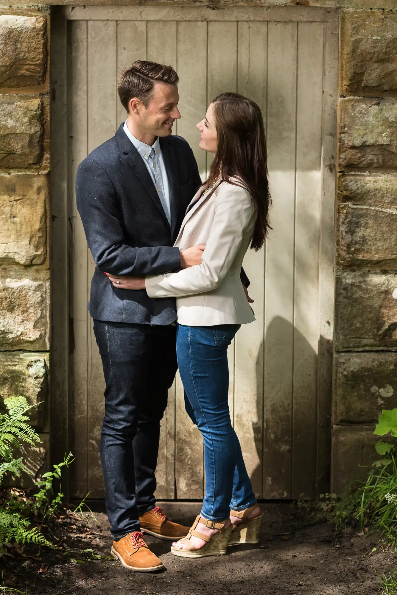 A couple gazing affectionately at each other, standing by a stone wall and wooden door, the man in a suit and the woman in a blazer and jeans.
