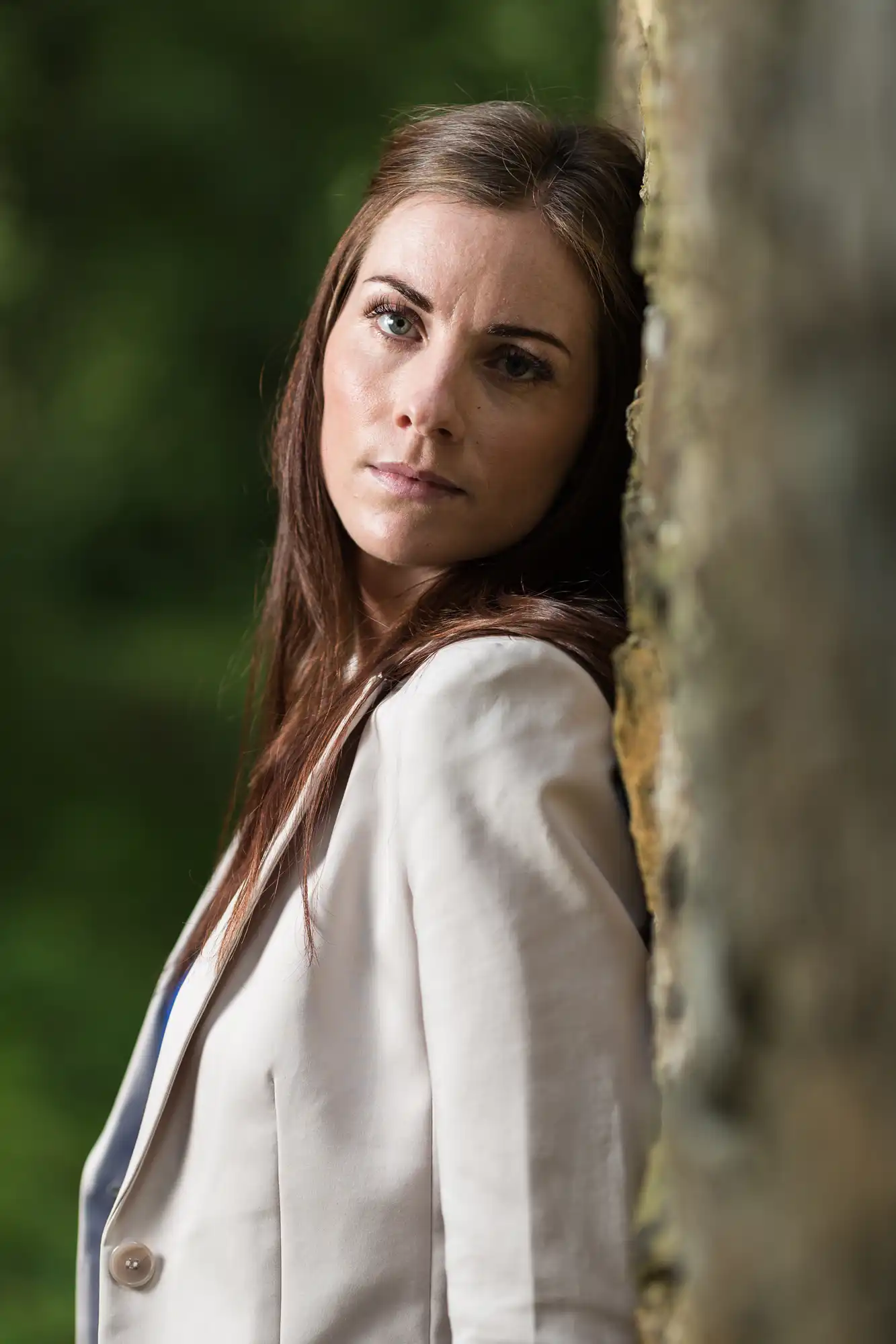 A woman with long brown hair, wearing a white blazer, looking thoughtfully past the camera while leaning against a tree in a wooded area.