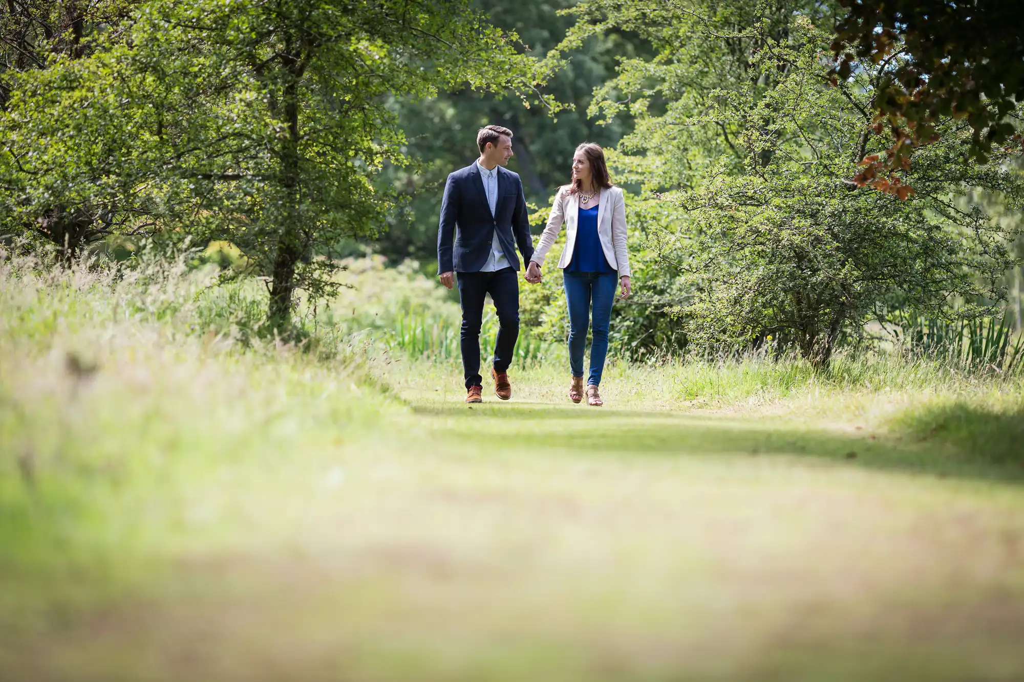 A young couple holding hands while walking on a sunny forest path. both are casually dressed, the man in a blazer and the woman in a blouse.
