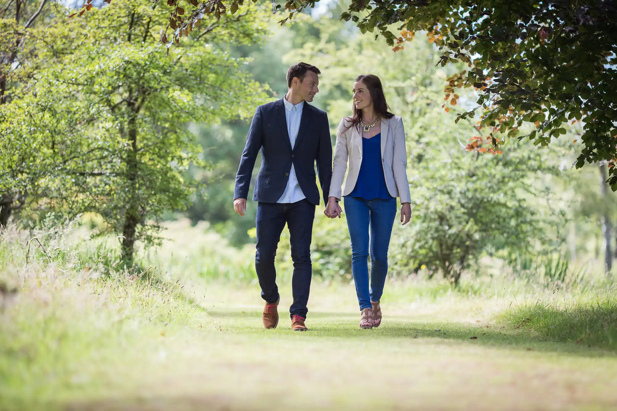 A couple walks hand in hand along a tree-lined path in a sunny park, both dressed in smart-casual attire.