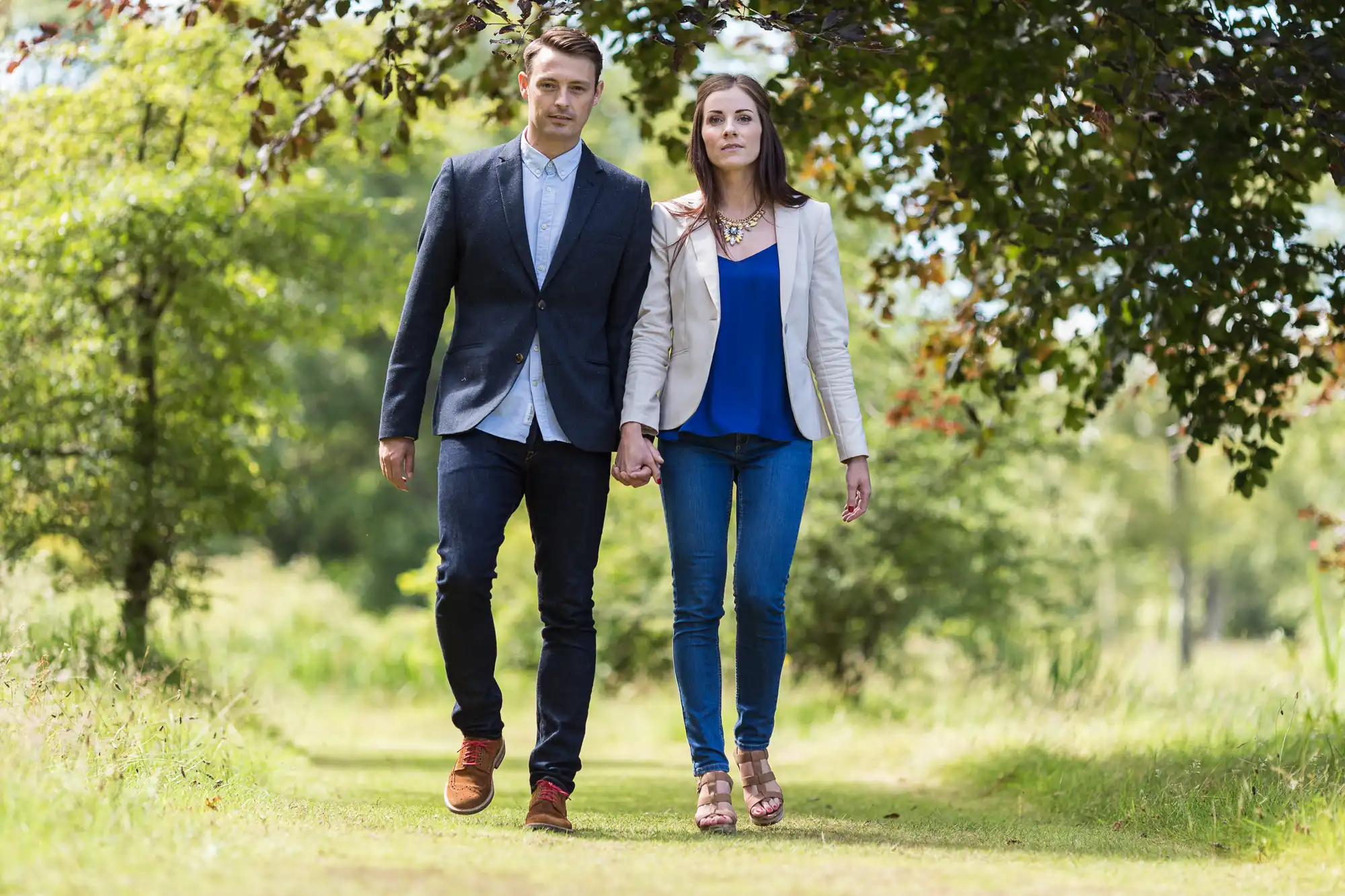 A young couple holding hands while walking down a sunny, tree-lined path. the man is wearing a blazer and jeans, and the woman is dressed in a blazer and jeans.
