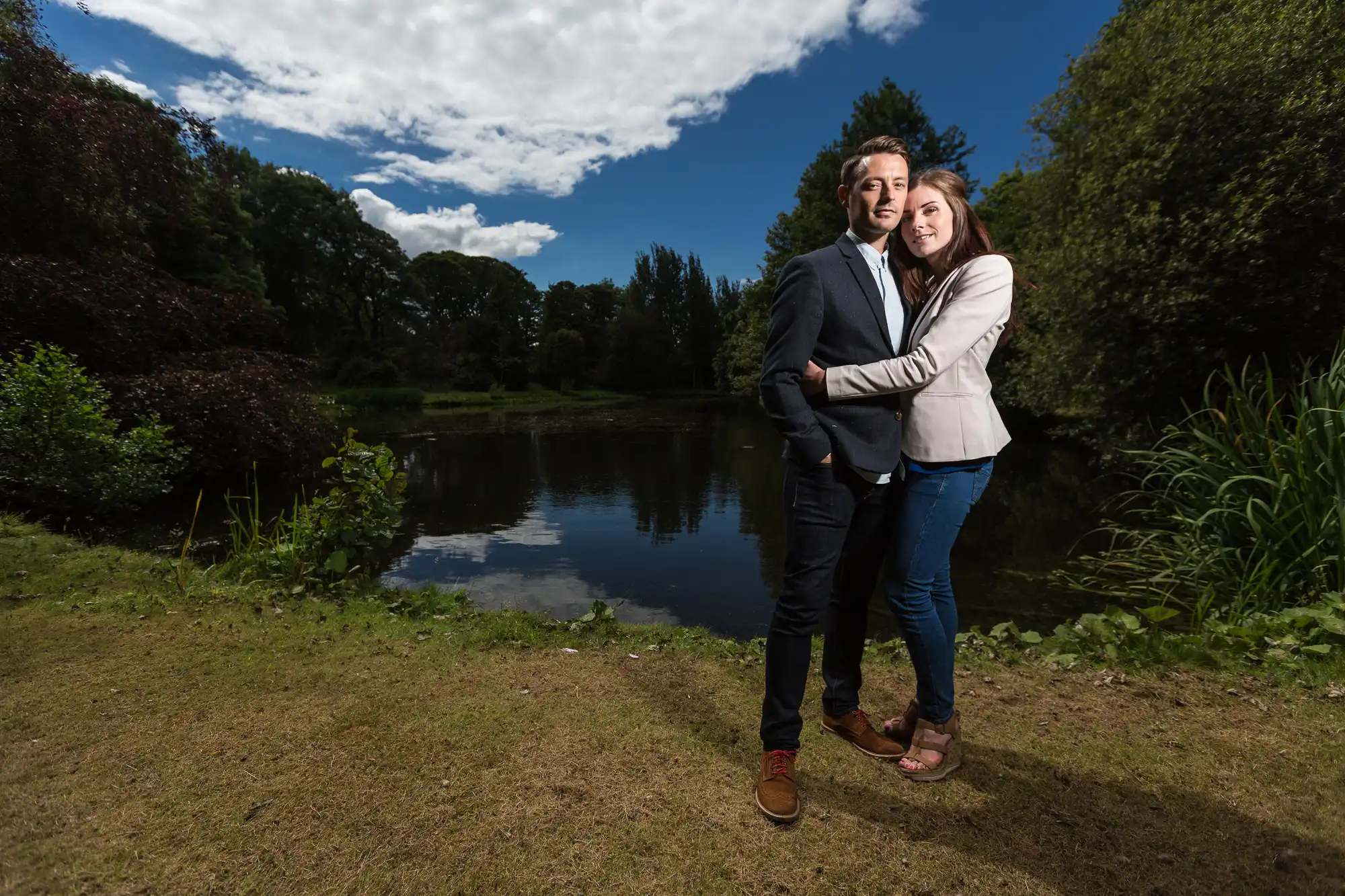 A couple embracing by a pond in a lush park, with clear blue skies and vibrant greenery surrounding them.