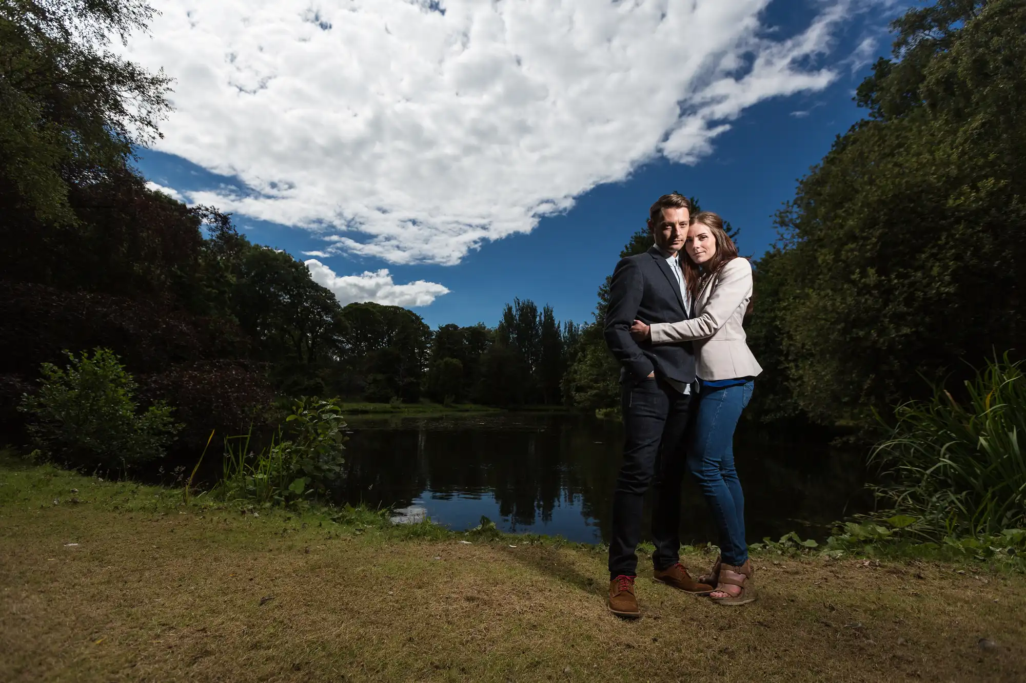 A couple embracing by a serene pond under a cloudy sky in a lush park.